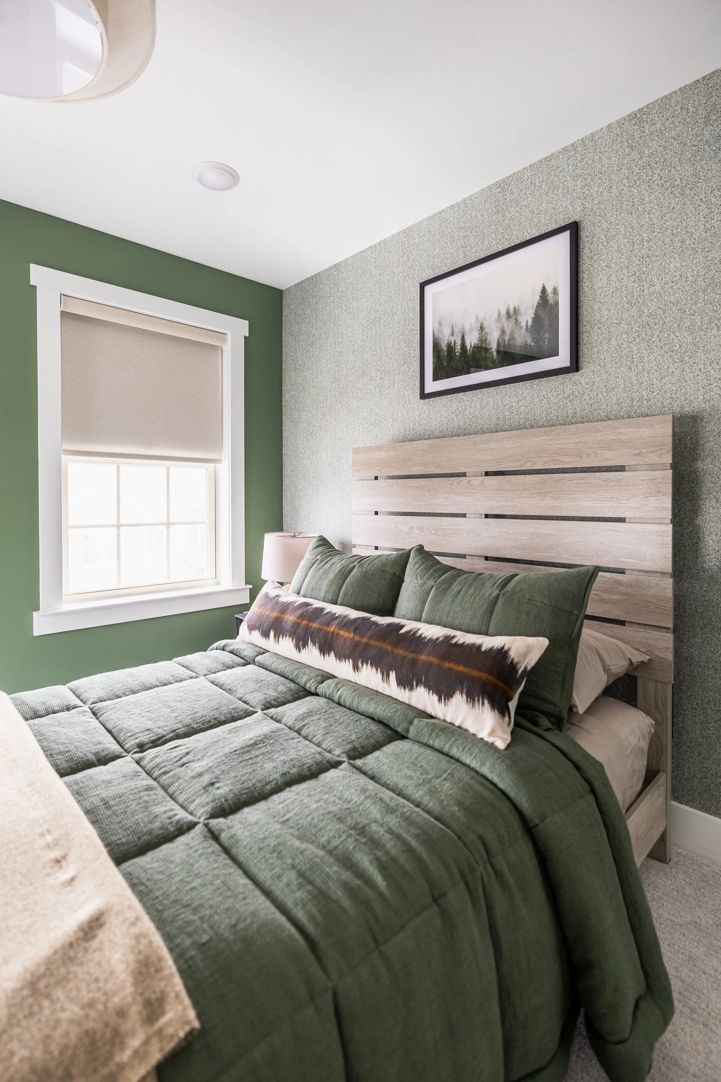 Modern bedroom with green walls and bedding, wooden headboard, and framed forest artwork above the bed. Window with beige shade on the left.