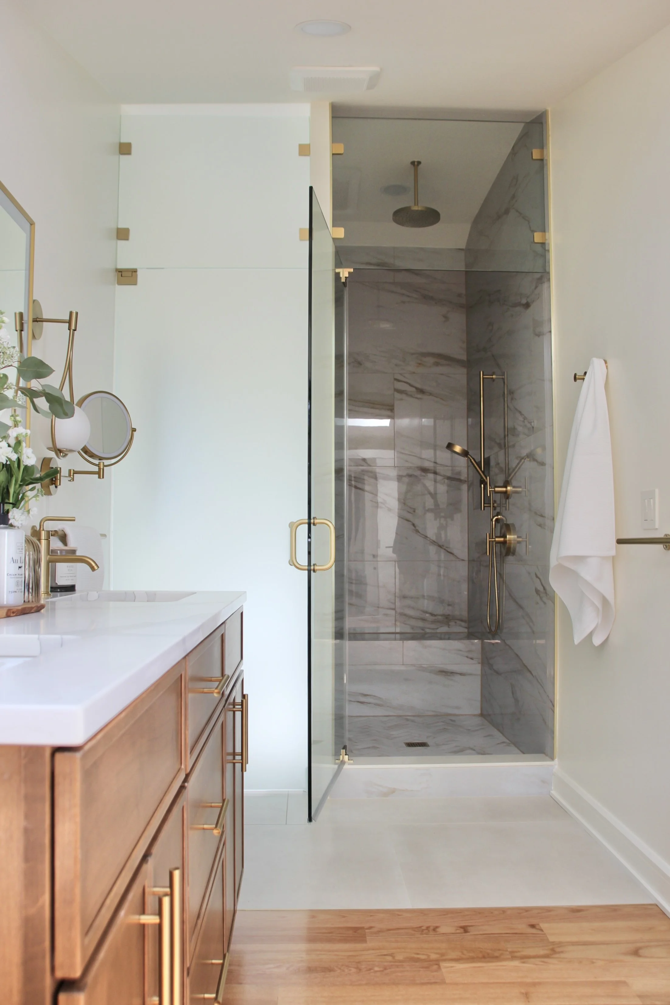 Modern bathroom with wood cabinetry, white countertop, gold fixtures, and a glass-enclosed shower with marble tiles.