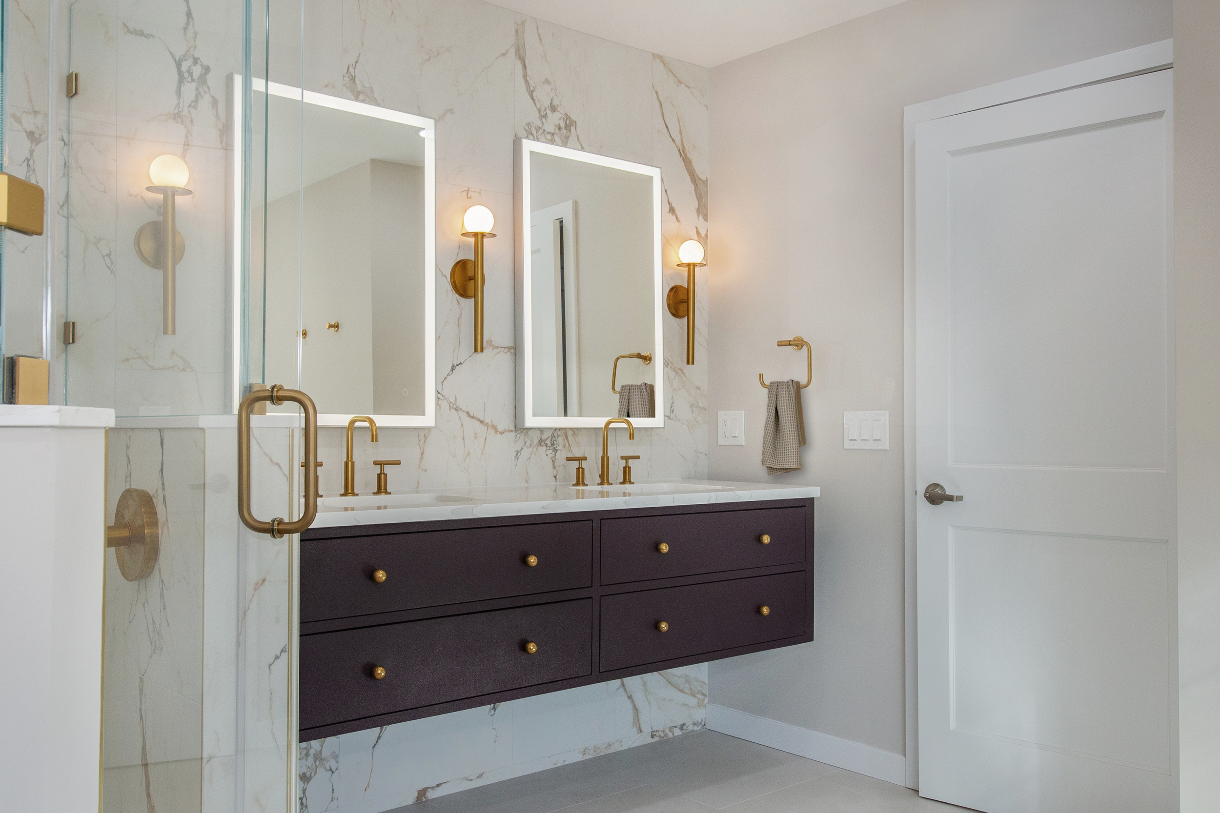 Modern bathroom with marble walls, double sink with dark vanity, gold fixtures, lighted mirrors, and glass shower.