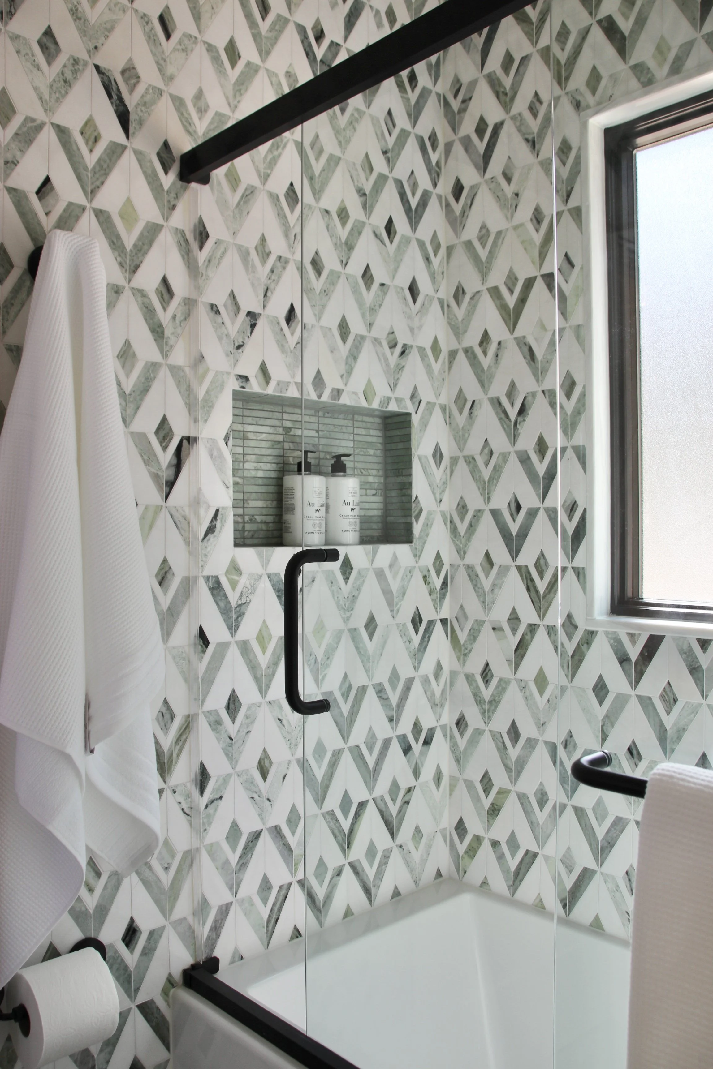 Modern bathroom with geometric patterned tiles, glass shower door, white towels, and window.