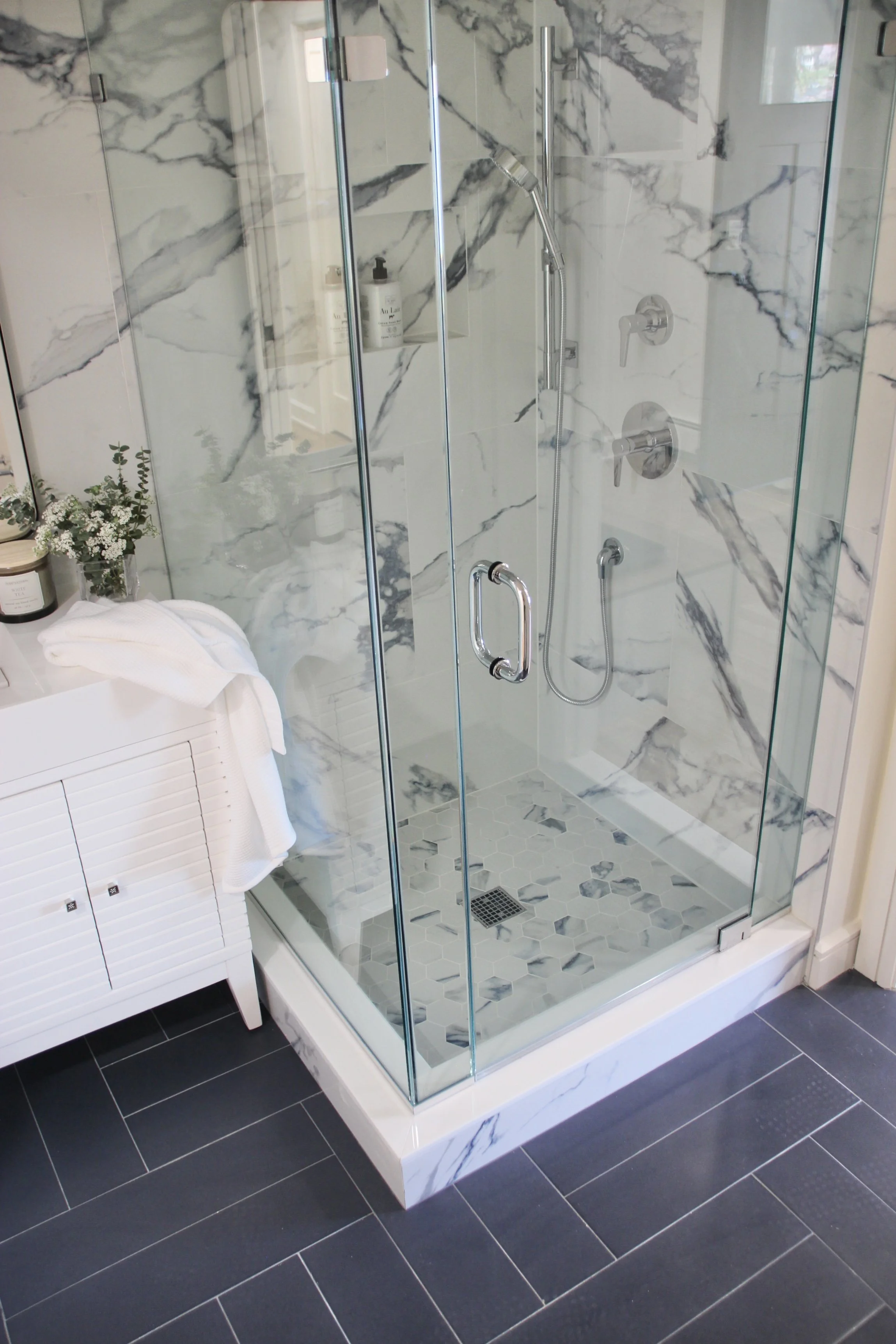 Modern bathroom with glass shower enclosure, marble wall tiles, hexagonal floor tiles, white vanity with towel, and dark tile flooring.