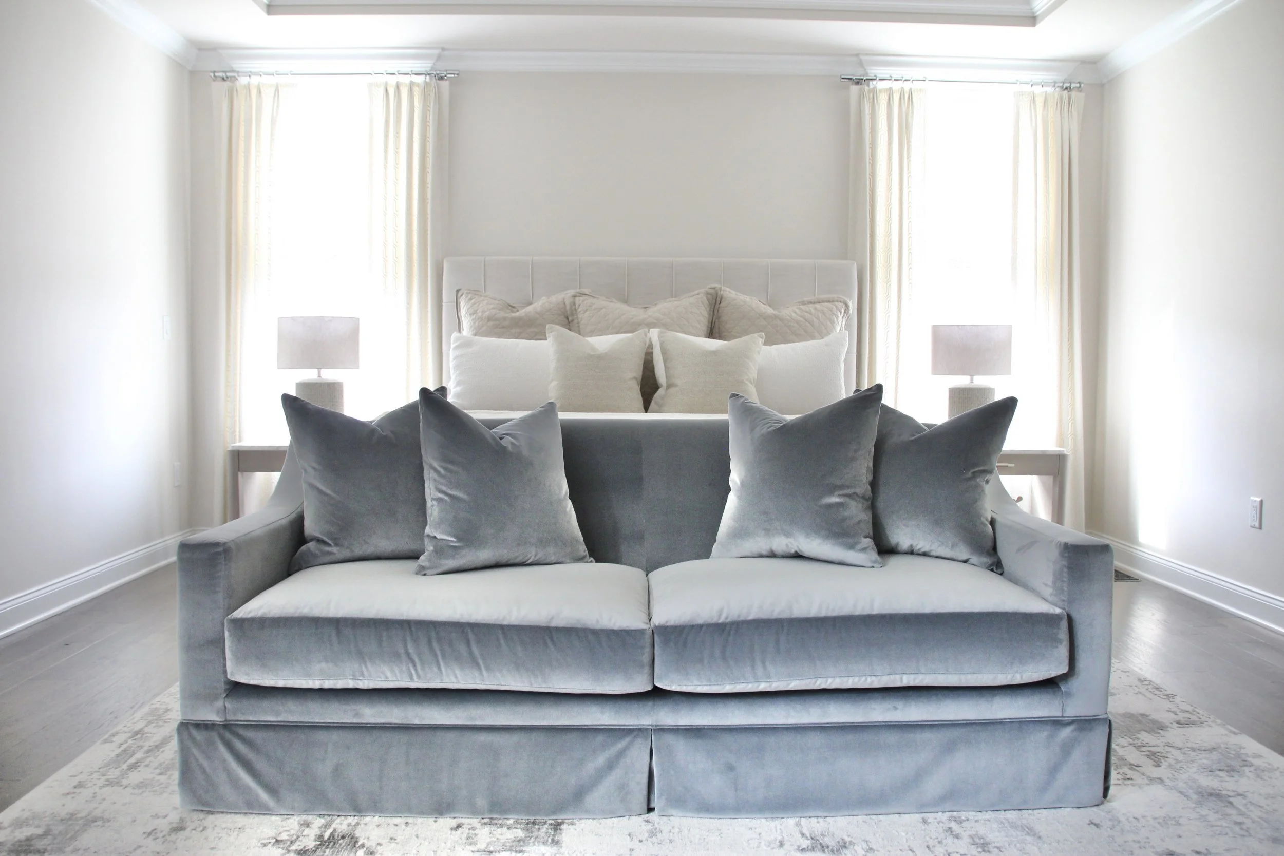 Bedroom with a gray sofa, white bed with multiple pillows, two side tables with lamps, and sheer curtains.