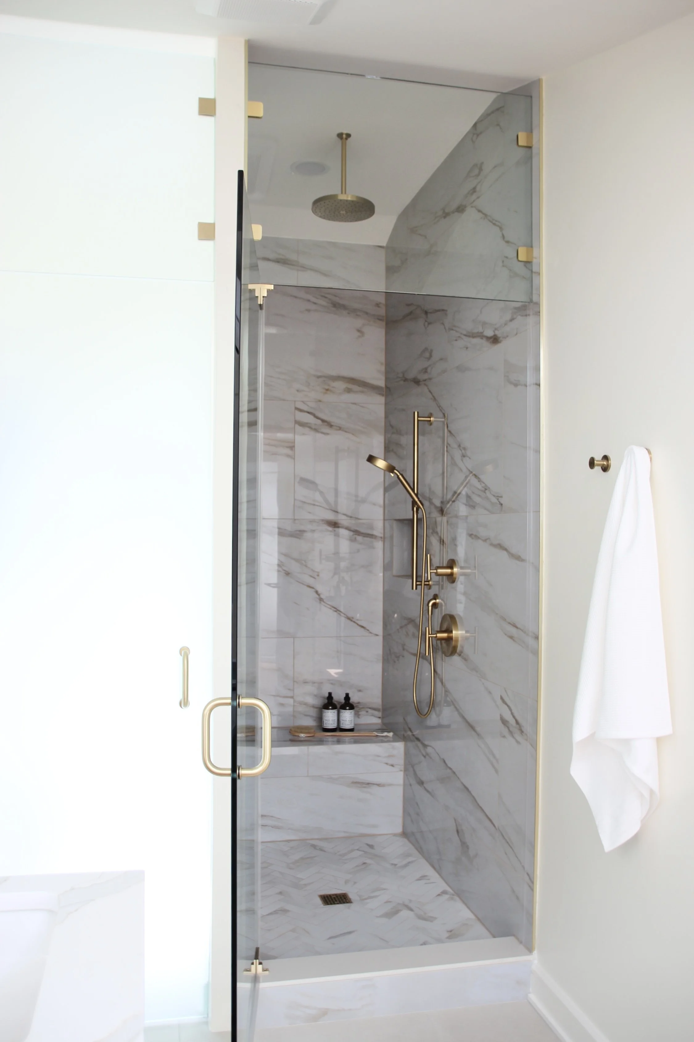 Modern bathroom with a marble shower, gold fixtures, and a glass door. A white towel is hanging on the wall, and shampoo bottles are on a shelf inside.