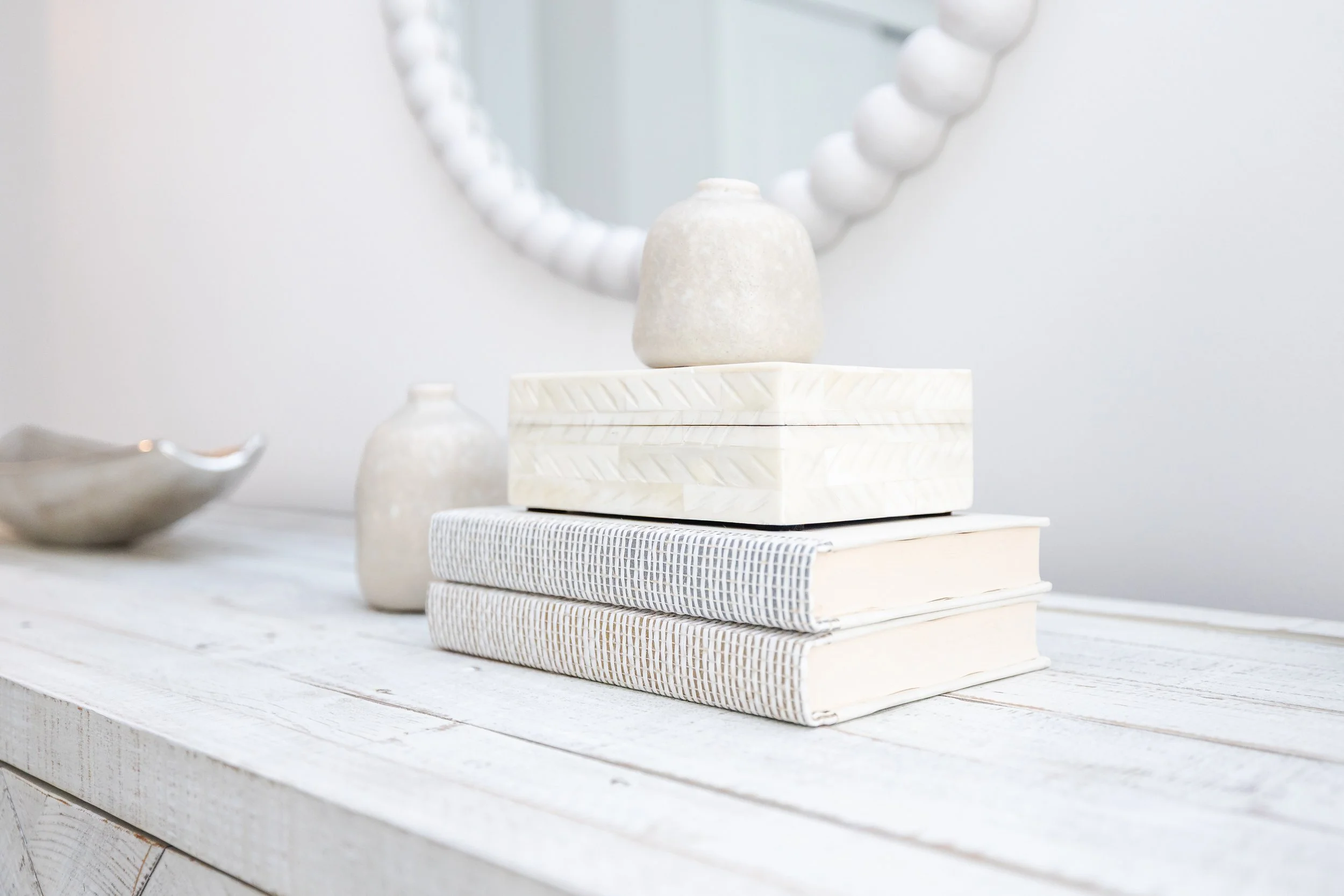 White rustic table with stacked books, small vases, and decorative box, mirror in background.