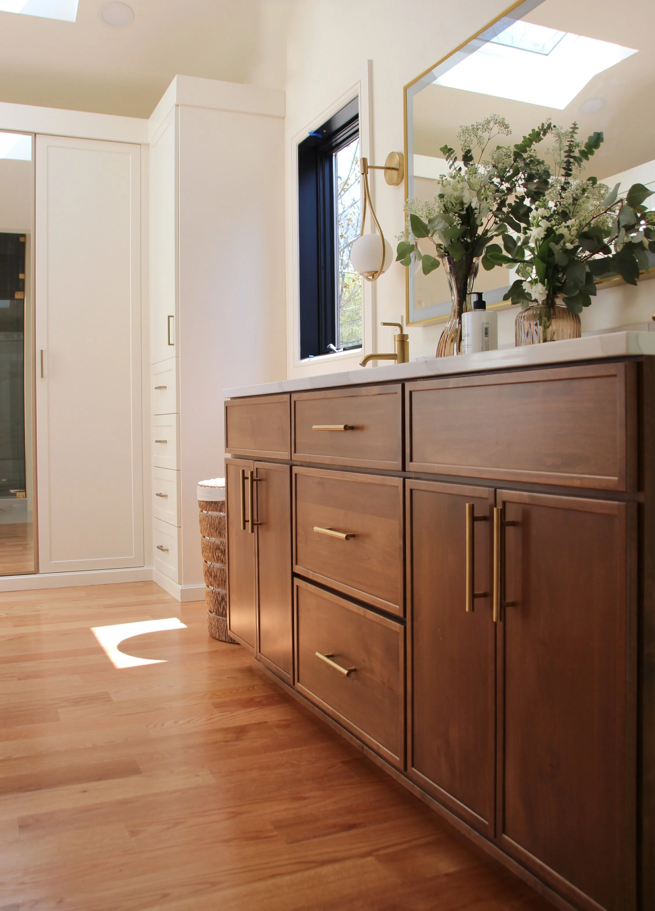 Modern bathroom with wooden vanity, brass fixtures, and floral decorations.