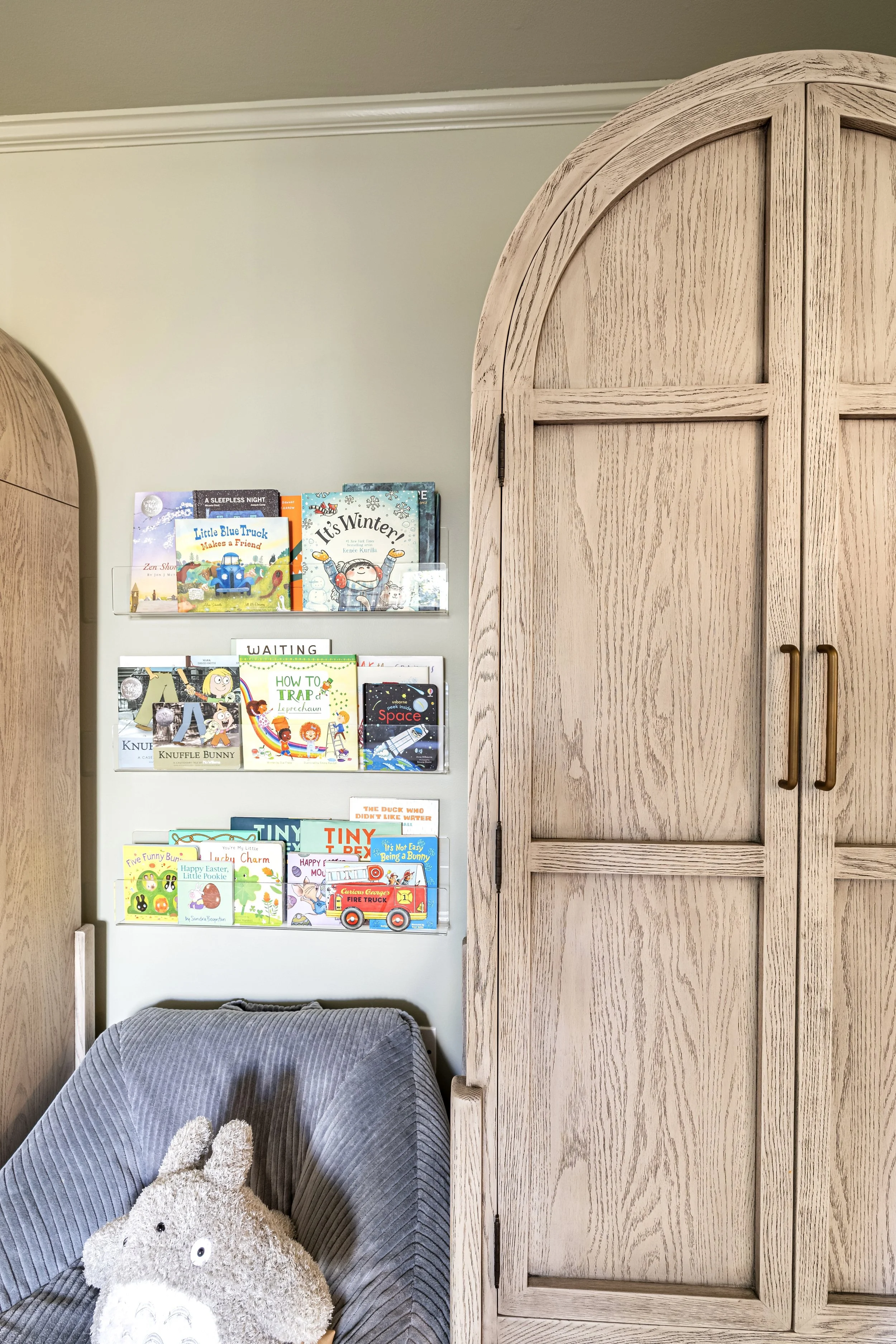 Children's books on wall shelves next to a light wood armoire in a room with a gray chair and plush toy.