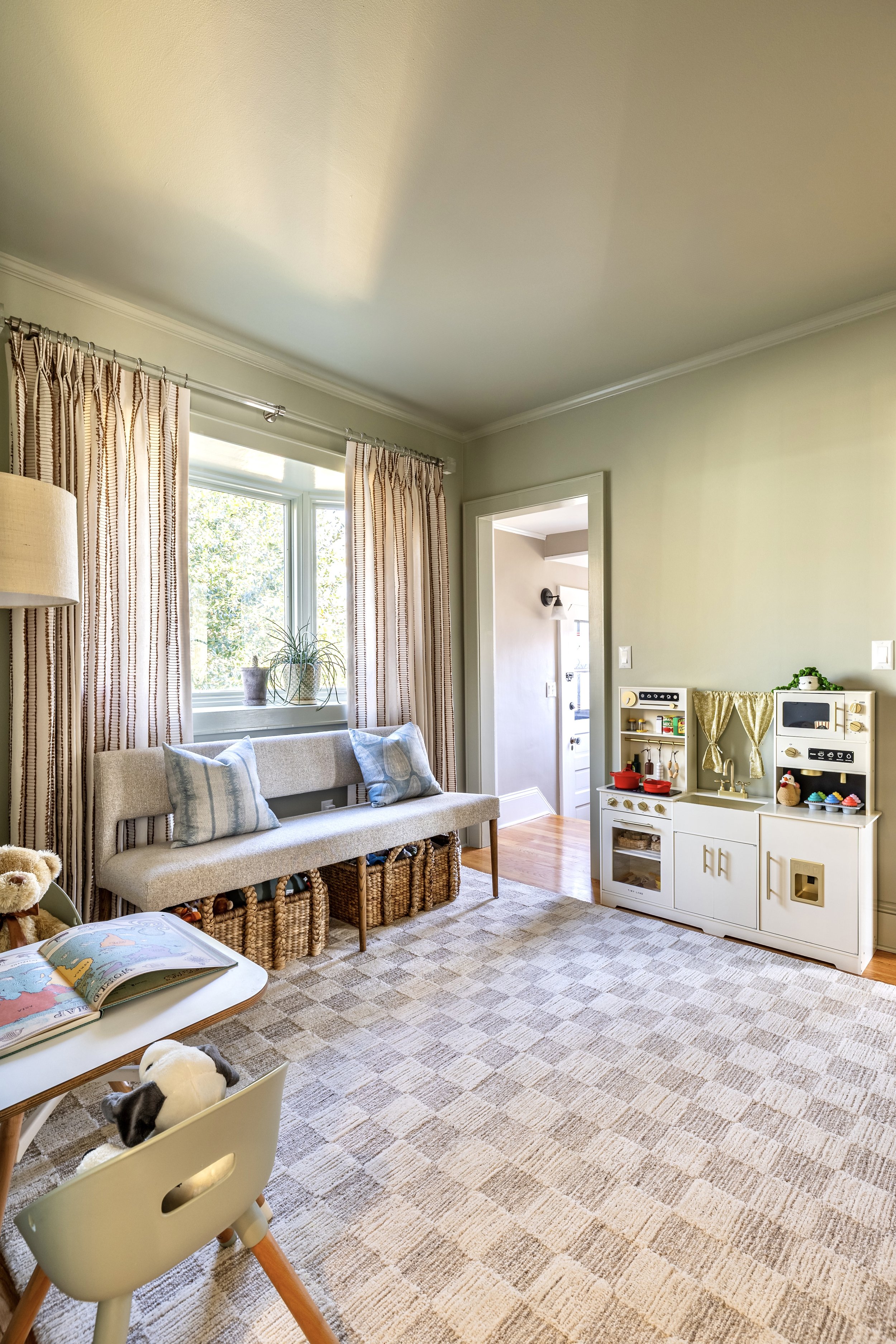 Children's playroom with a beige checkered rug, toy kitchen set, and bench with cushions. Wooden baskets are under the bench. A window with light curtains is in the background.