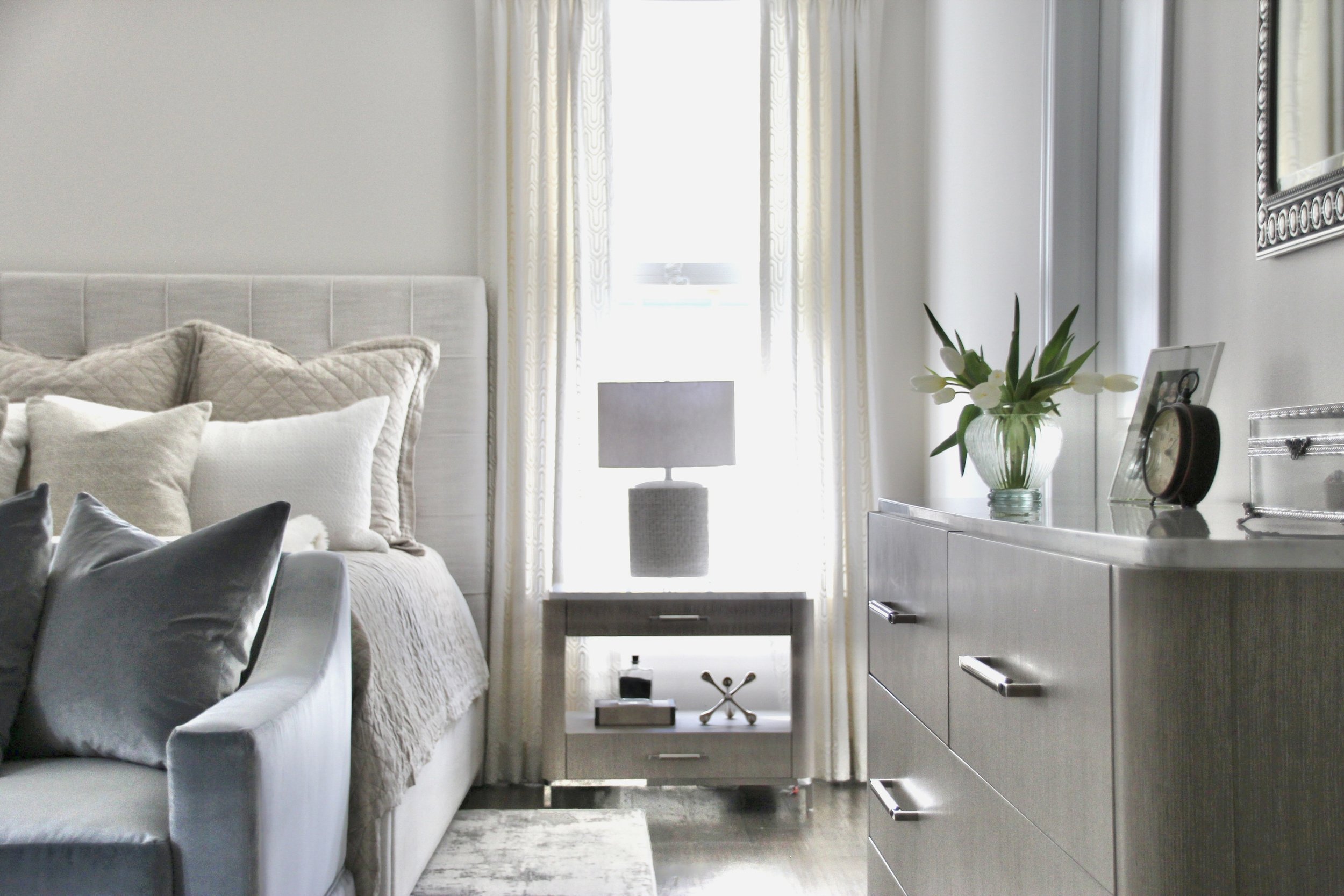 Modern bedroom with soft grey decor, featuring a bed with pillows, a nightstand with a lamp, and a dresser with a vase of flowers.