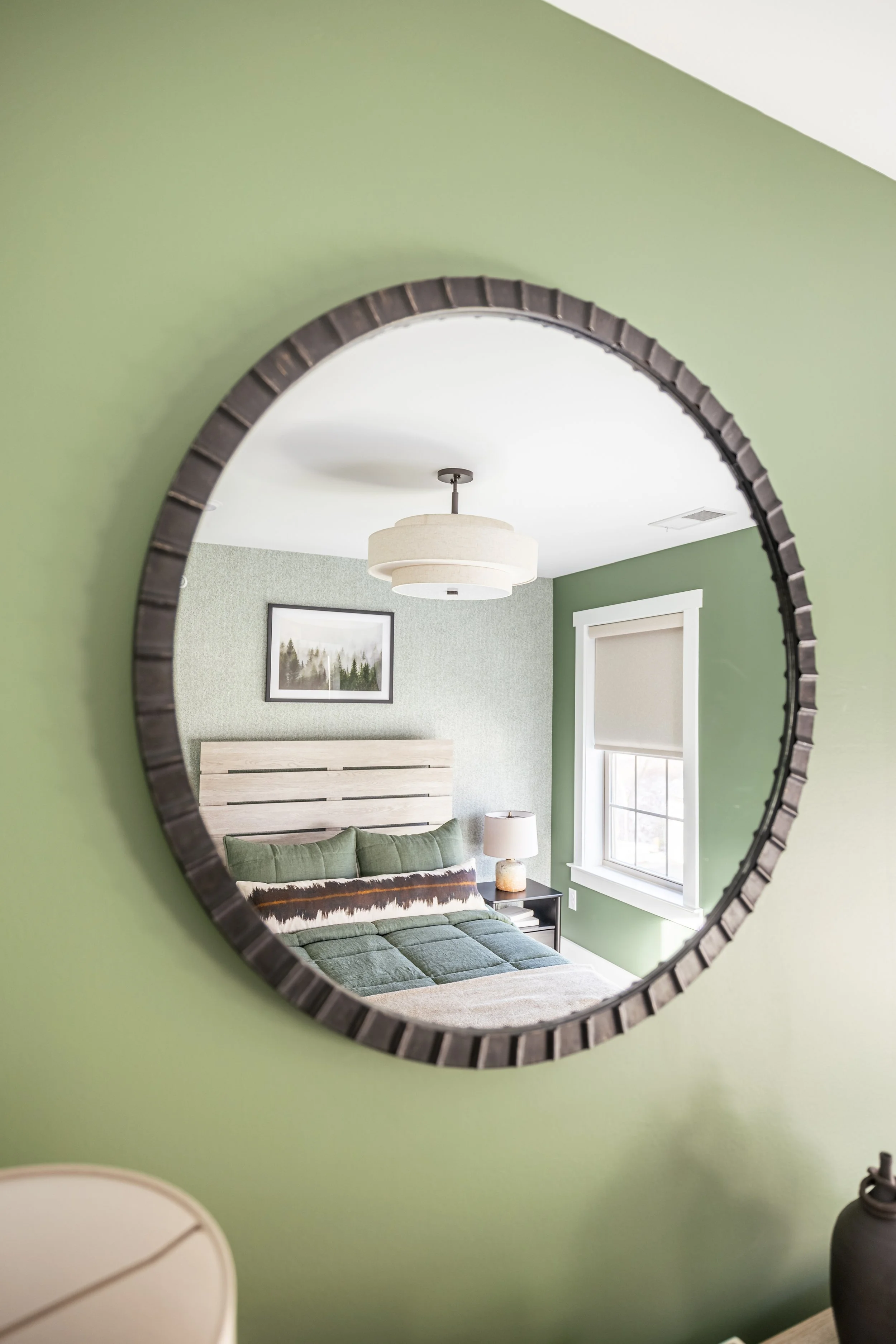 Reflection in circular mirror showing a modern bedroom with green walls, a wooden headboard, green bedding, a landscape picture, and a pendant light.