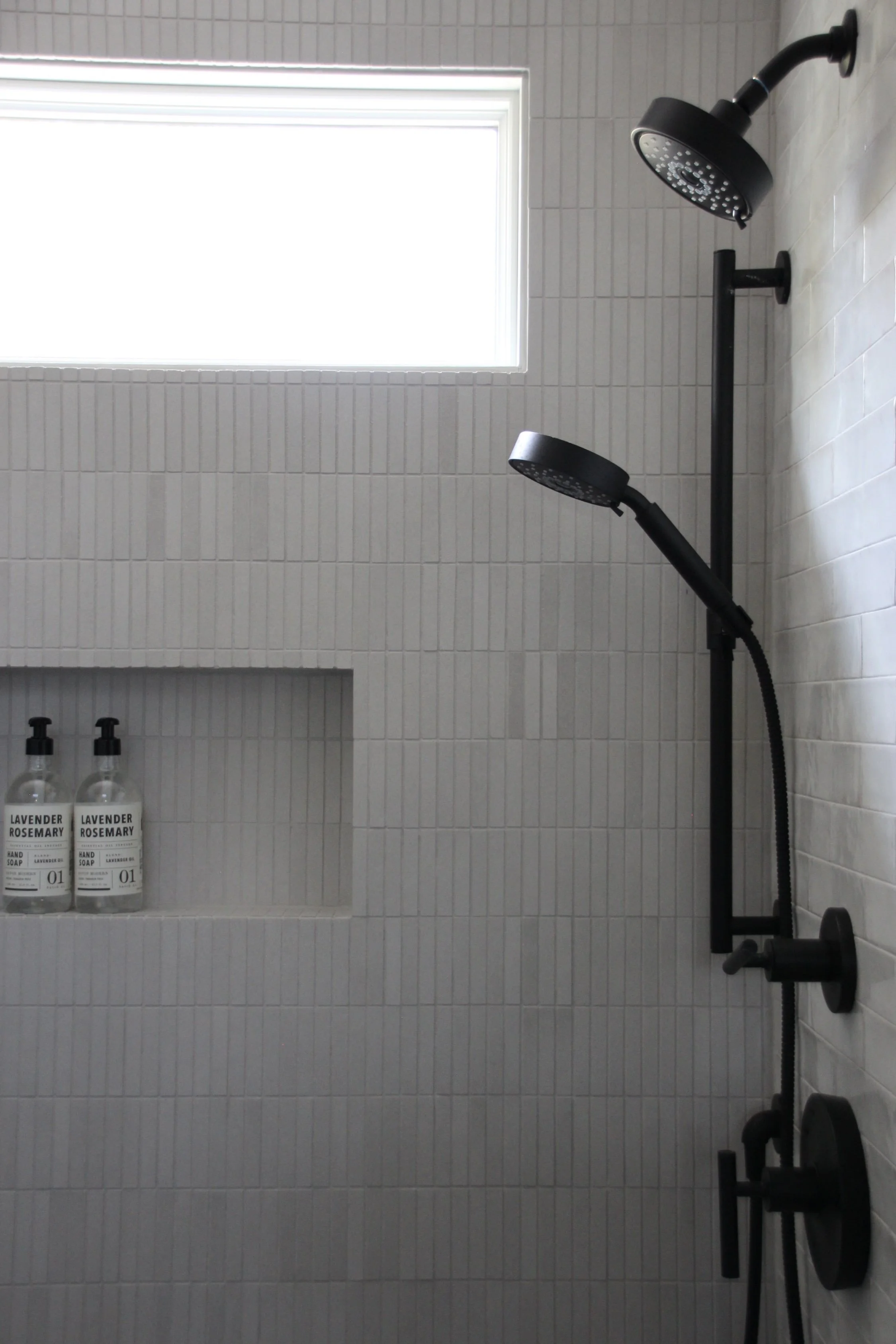 Modern shower with black fixtures, handheld showerhead, overhead shower, and built-in shelf with two bottles of soap. Vertical white tiles and rectangular window.