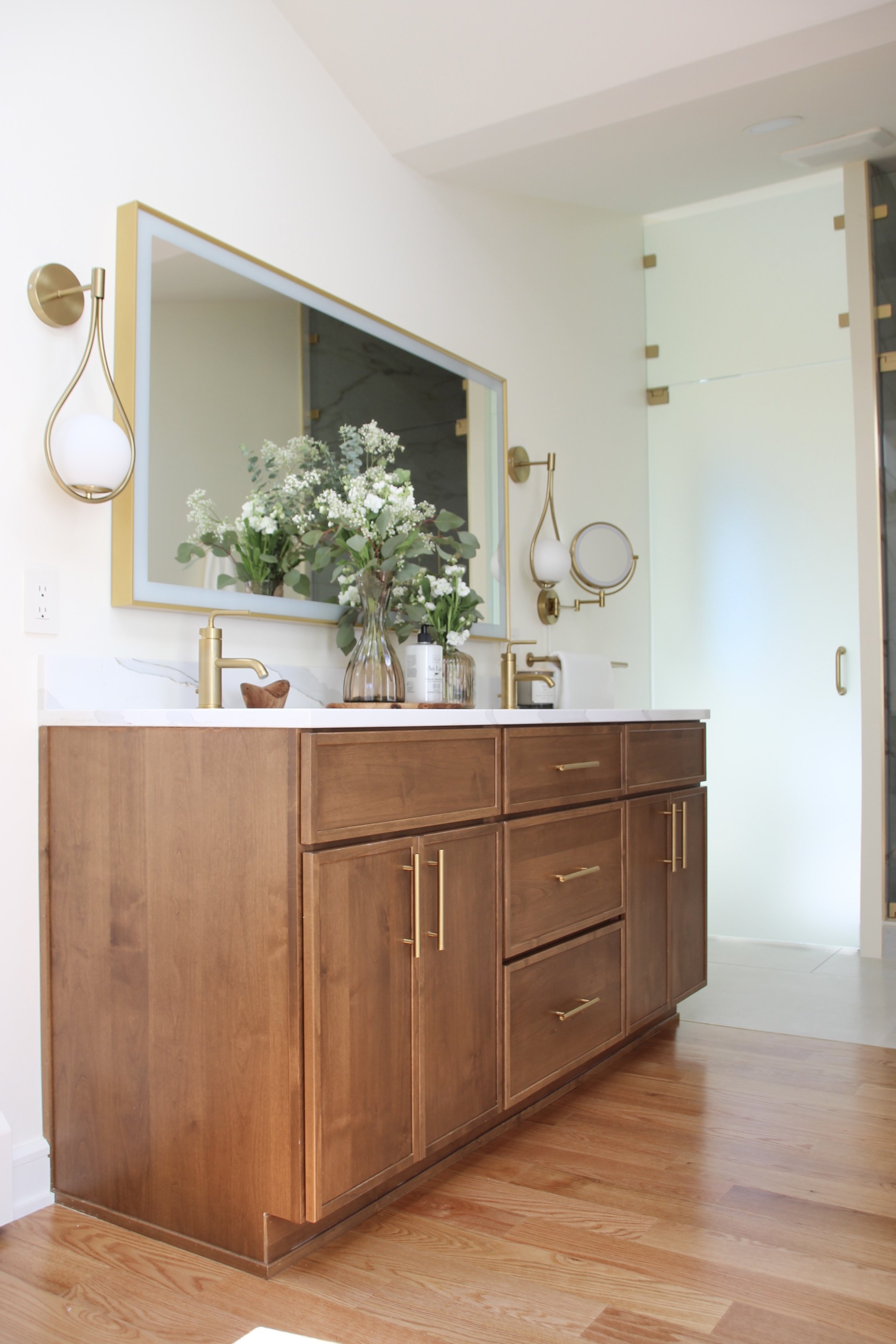 Modern bathroom with wood vanity, gold accents, large mirror, and decorative lighting.