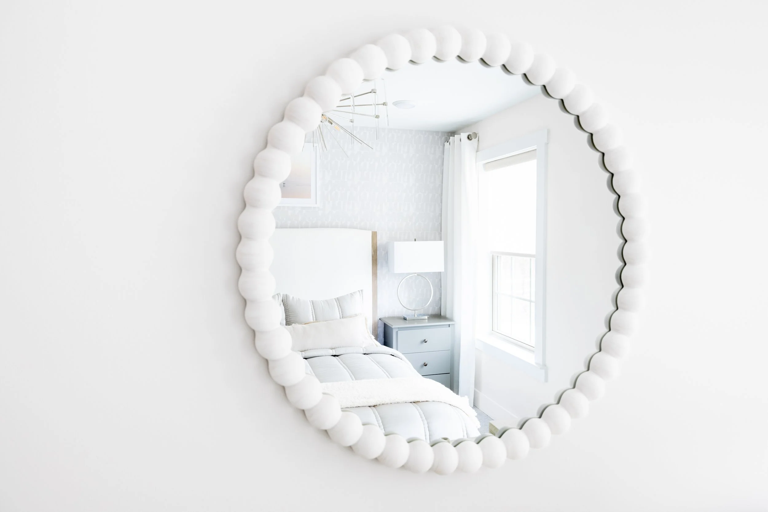 Round decorative mirror reflecting a modern bedroom with a bed, nightstand, lamp, and window.