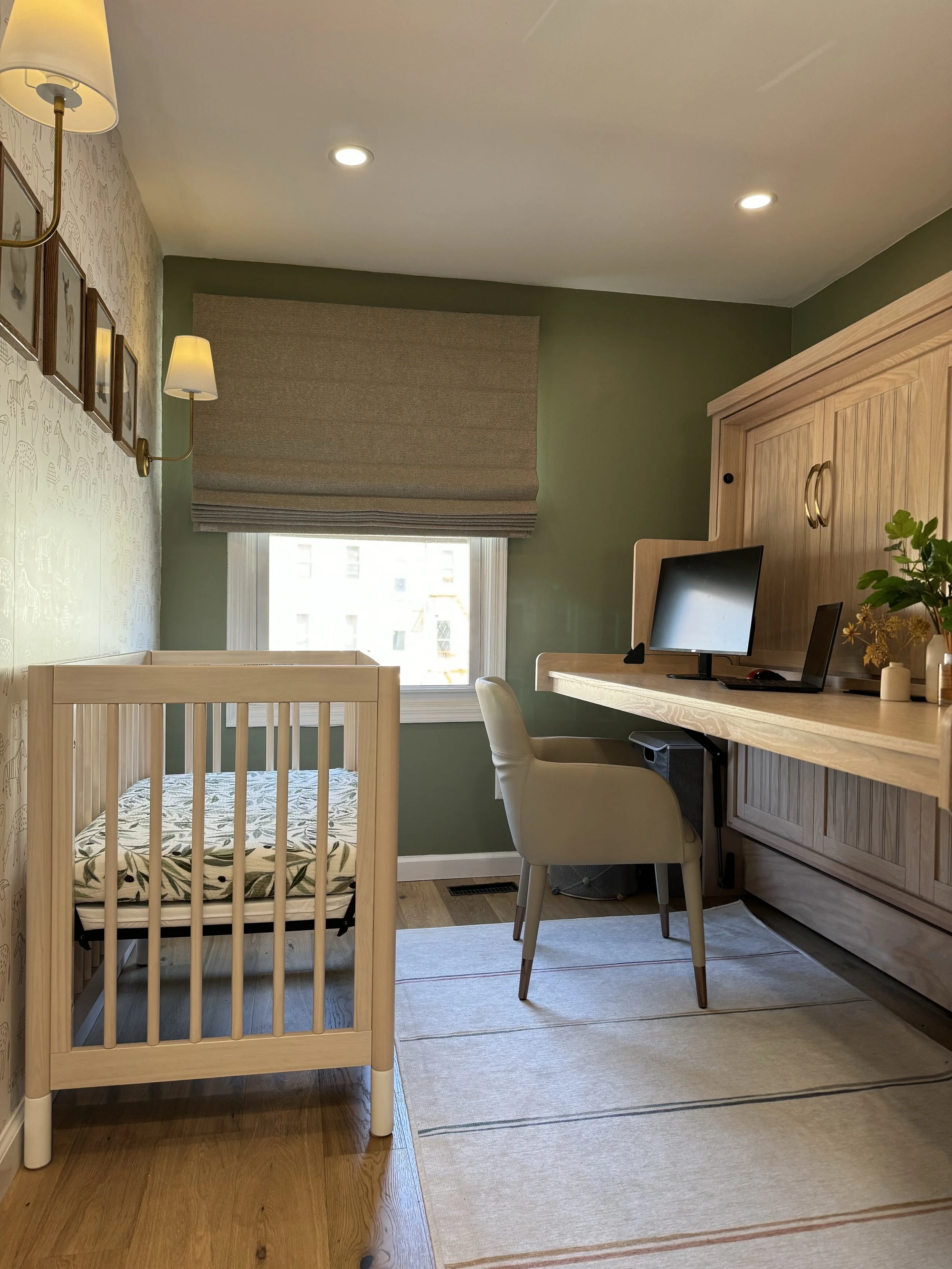 A small room featuring a crib with a patterned mattress on the left, a window with a Roman shade, a light wood desk with a computer monitor, and a modern chair. The walls are painted green, and there is decorative wall art. The floor is hardwood with