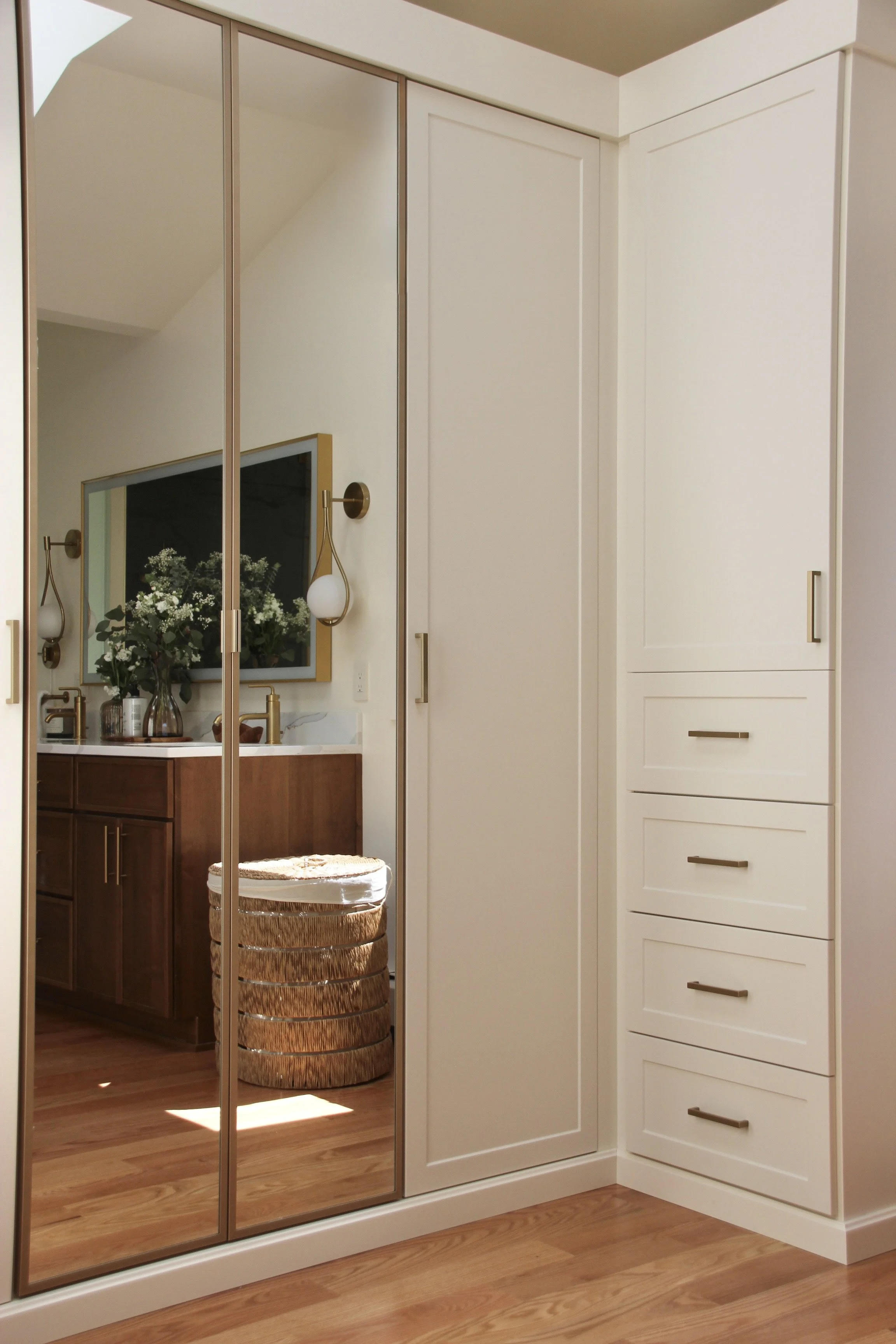 Modern bathroom with mirrored closet doors, white cabinetry with silver handles, wooden vanity with a countertop, wall sconces, and a woven laundry basket on wooden flooring.