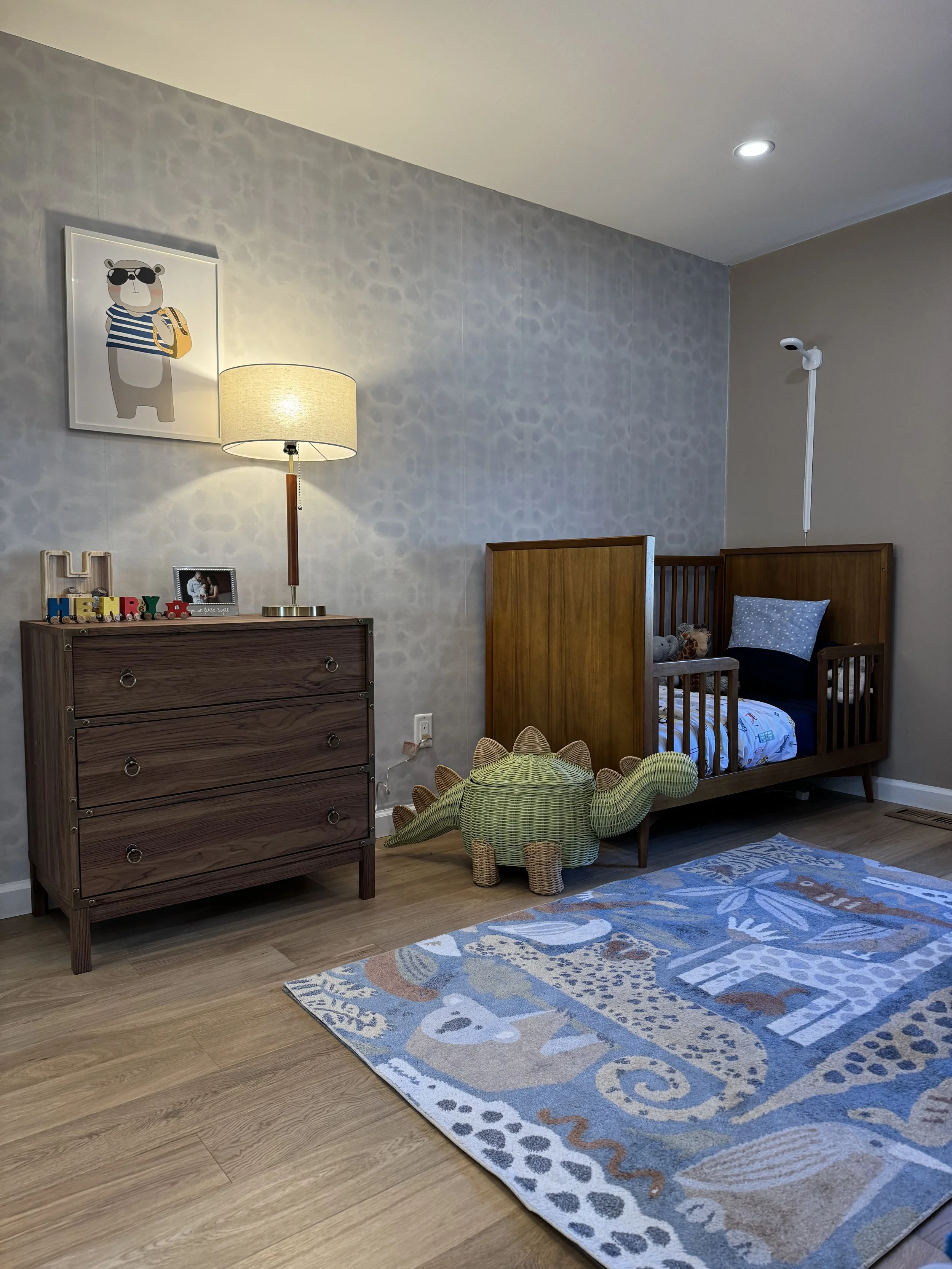 A child's room with a crib, dresser, and decorative items. The crib is wooden with blue bedding, and there's a plush animal toy inside. A dresser with drawers is next to it, with a lit lamp on top. A wall picture of a bear wearing sunglasses is above