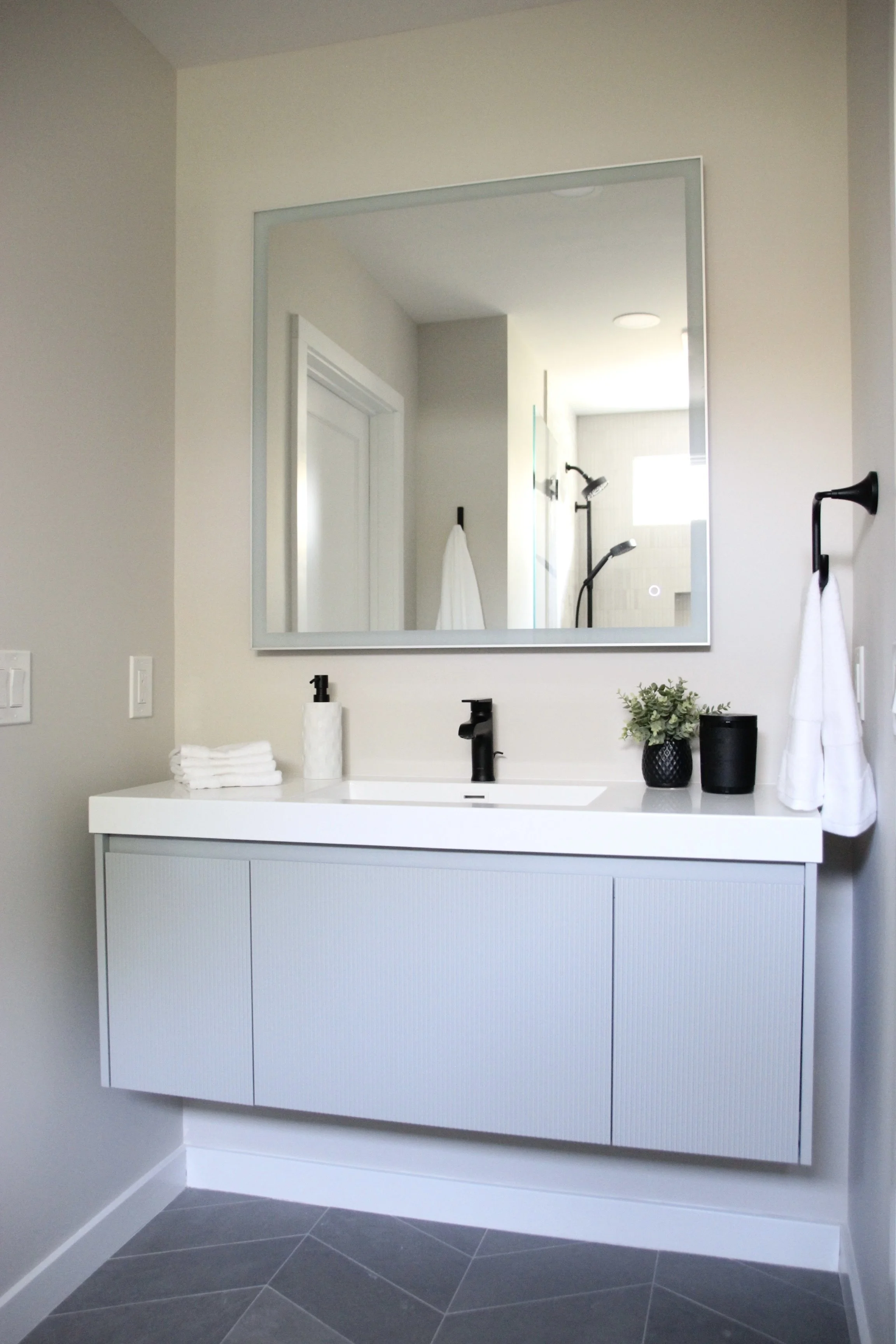 Modern bathroom with large mirror, a white floating vanity, black faucet, hand soap dispenser, rolled towels, potted plant, and wall-mounted towel rack.