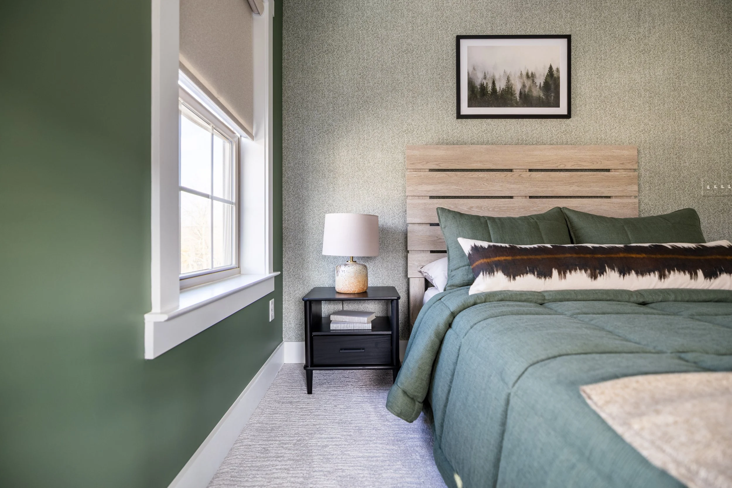 Modern bedroom with green walls, a bed with green bedding, a wooden headboard, and a decorative pillow. A nightstand with a lamp and a framed forest picture on the wall.
