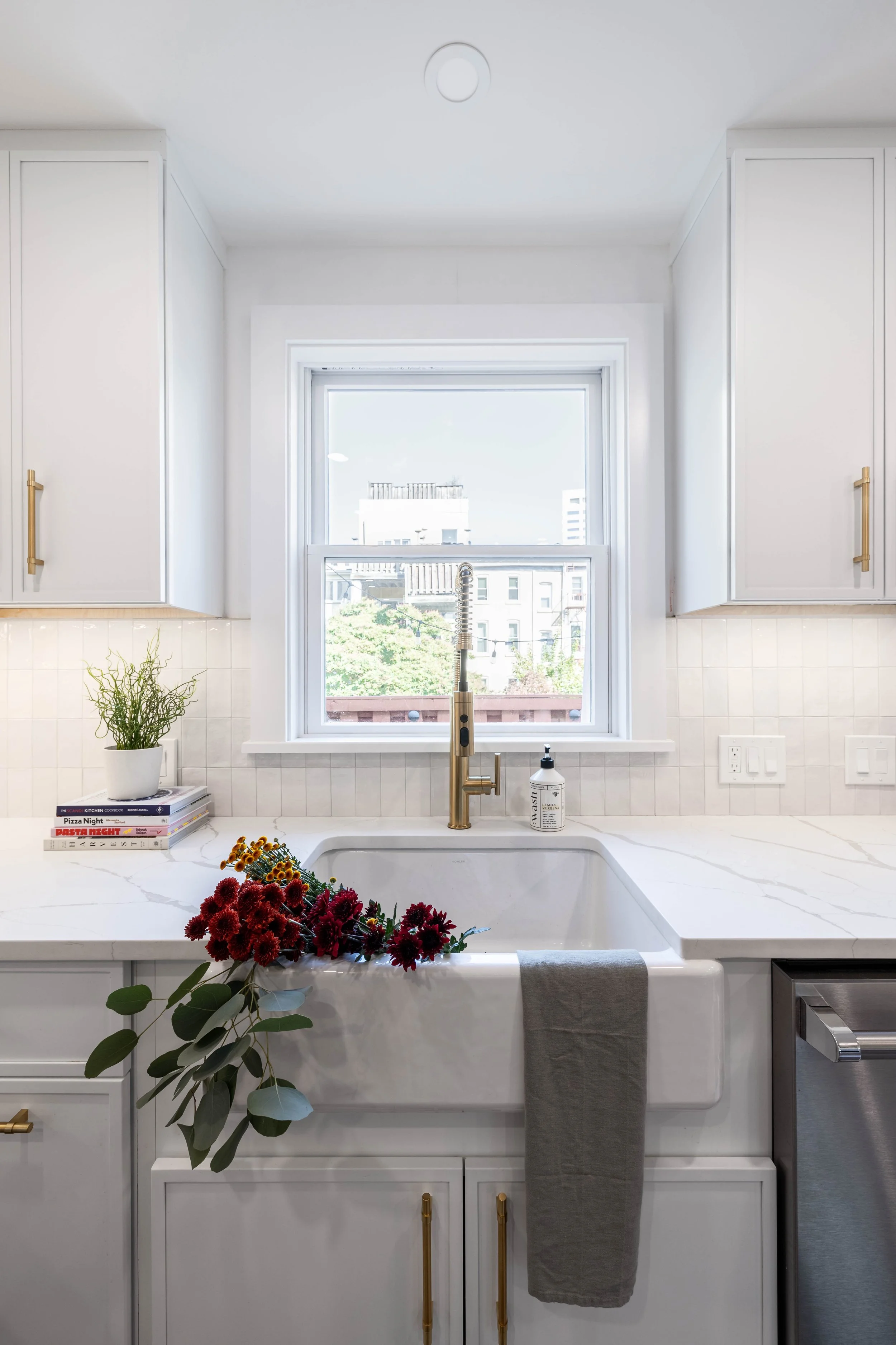 Modern kitchen sink with brass faucet, white cabinets, flowers on the countertop, and a window view.