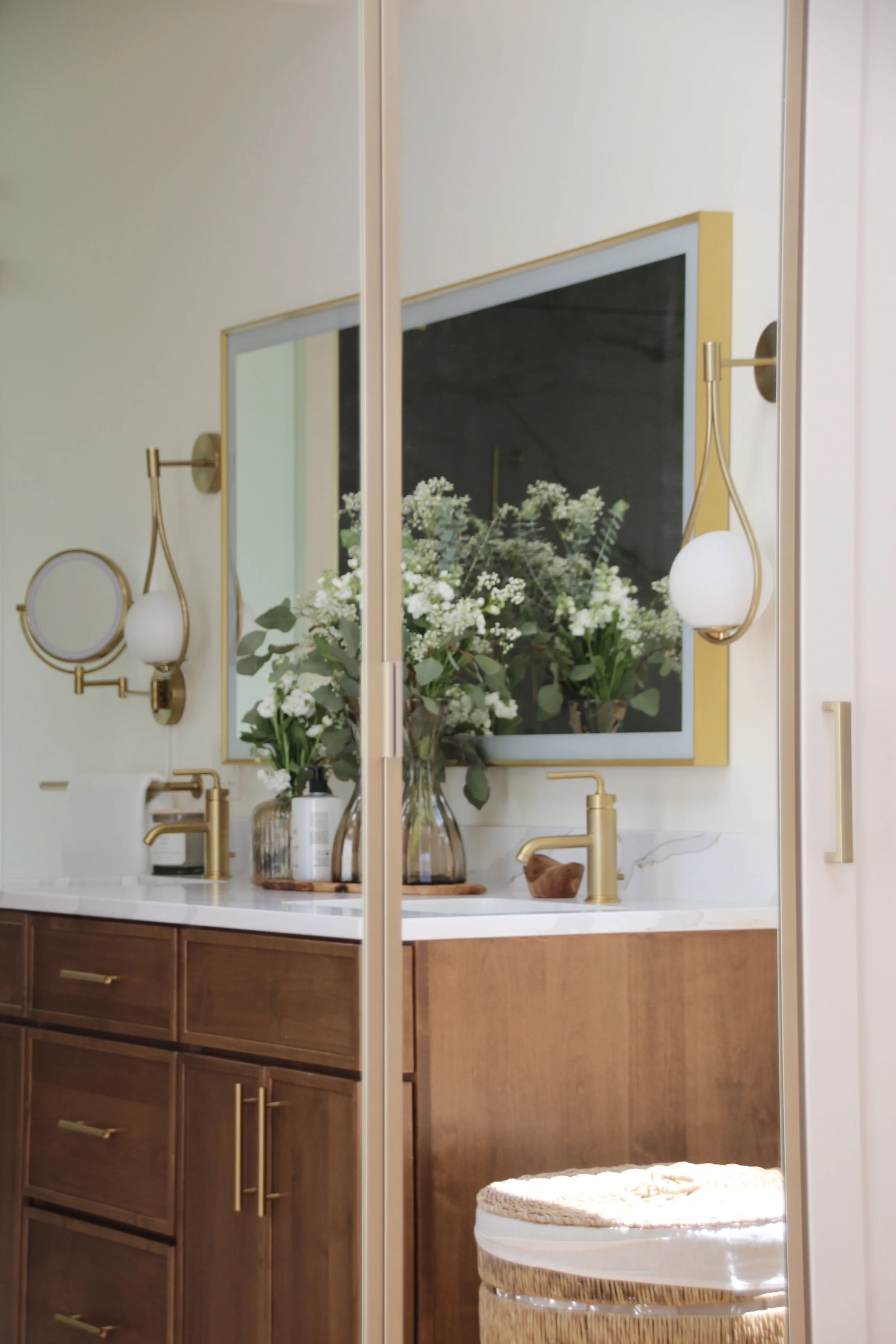 Modern bathroom with wooden cabinets, gold faucets, and mirror; decorative plants on counter, wicker basket visible.