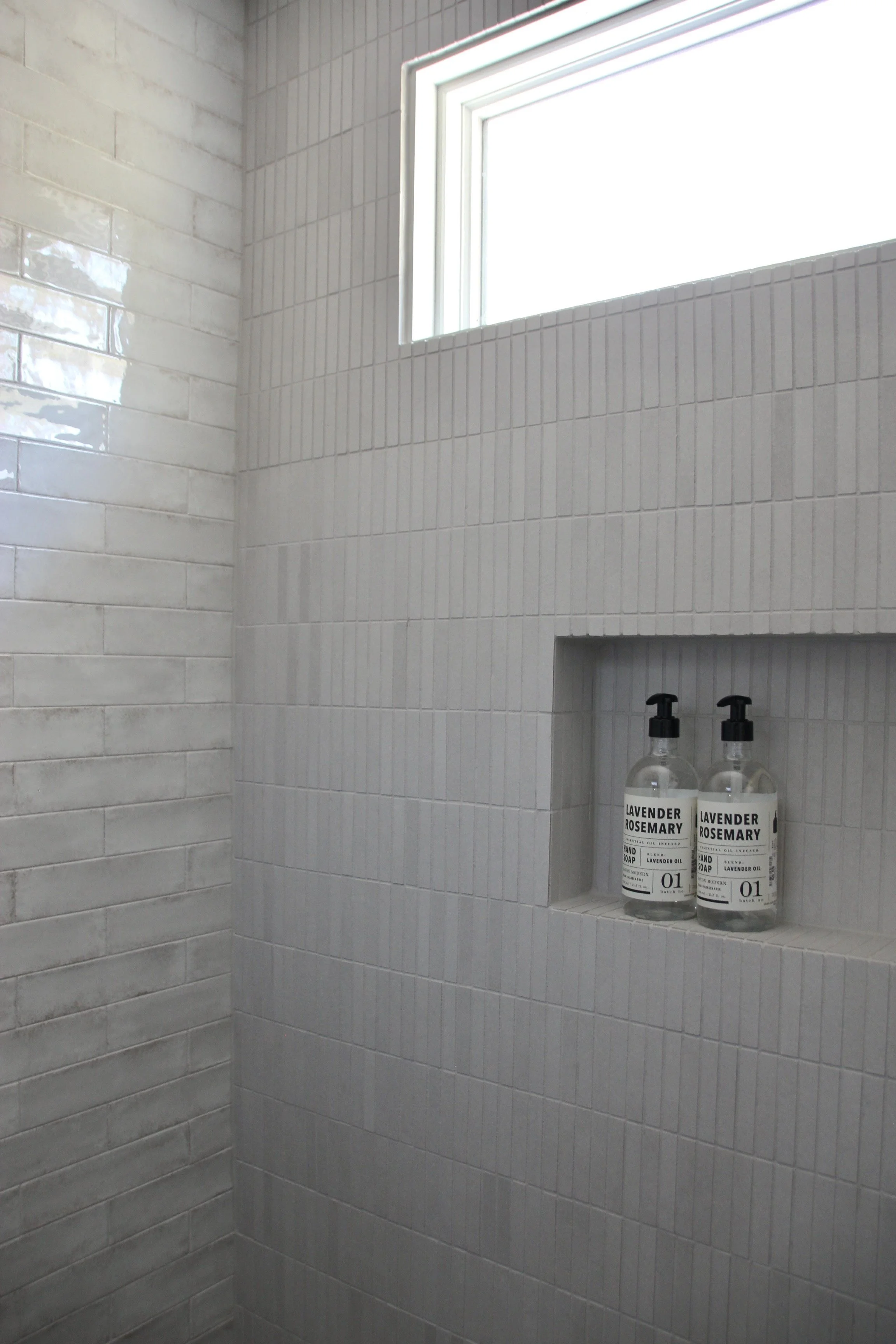 Bathroom shower with white subway tiles and a recessed shelf holding two bottles of lavender rosemary hand soap, lit by a small window.