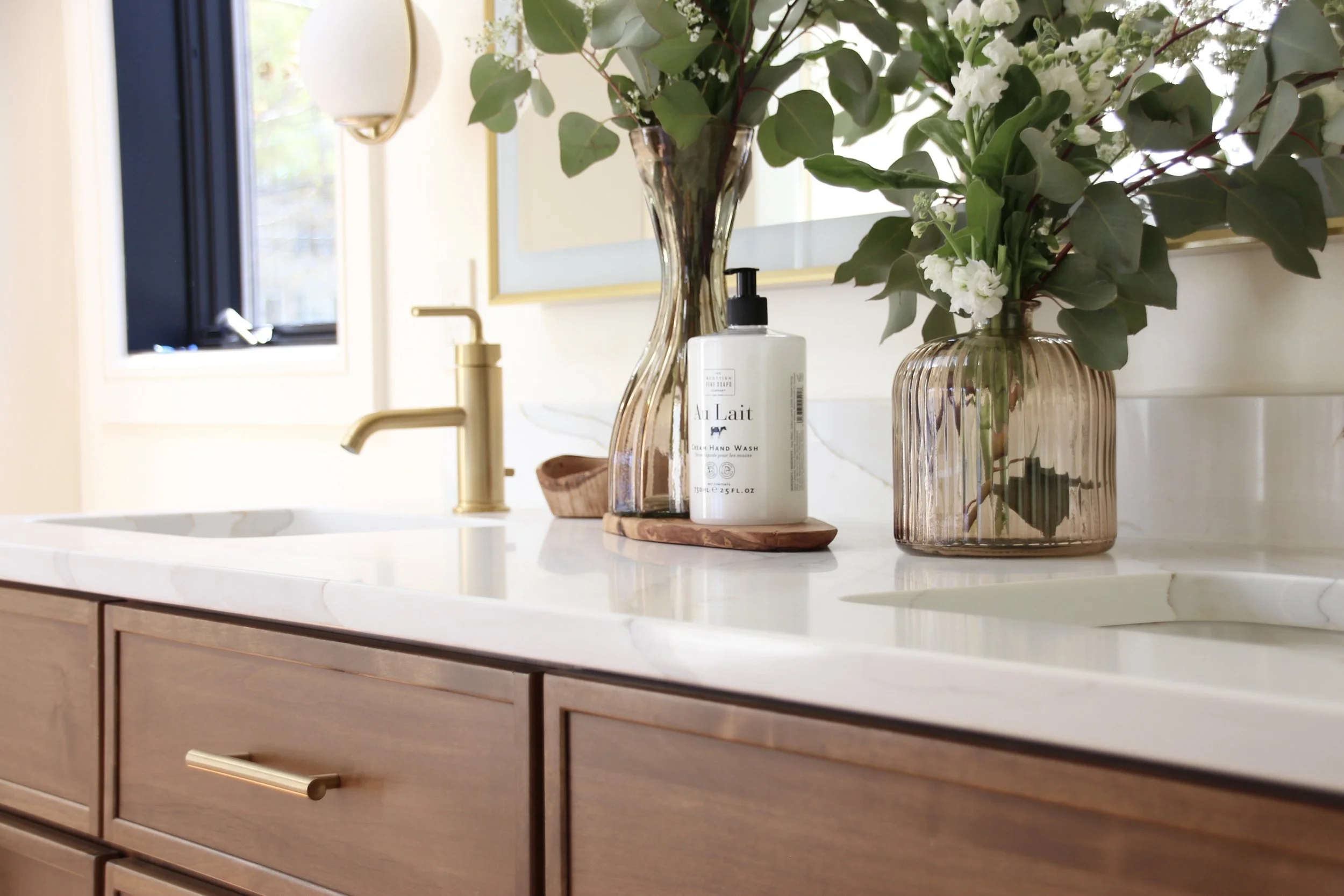 Bathroom vanity with gold faucet, flowers in vases, and hand wash bottle on marble countertop.