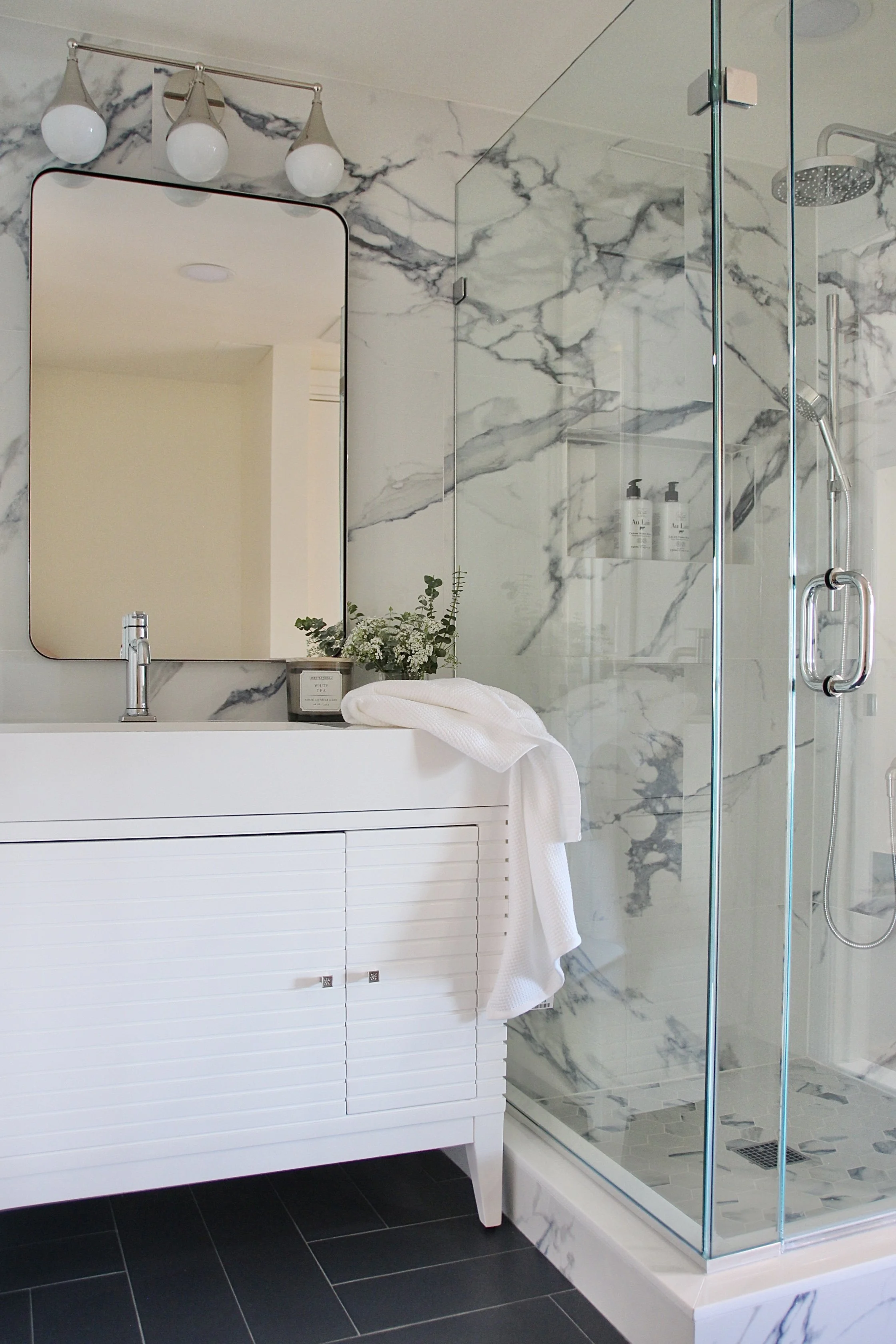 Modern bathroom with glass shower, marble walls, white vanity, mirror, and black tile floor.