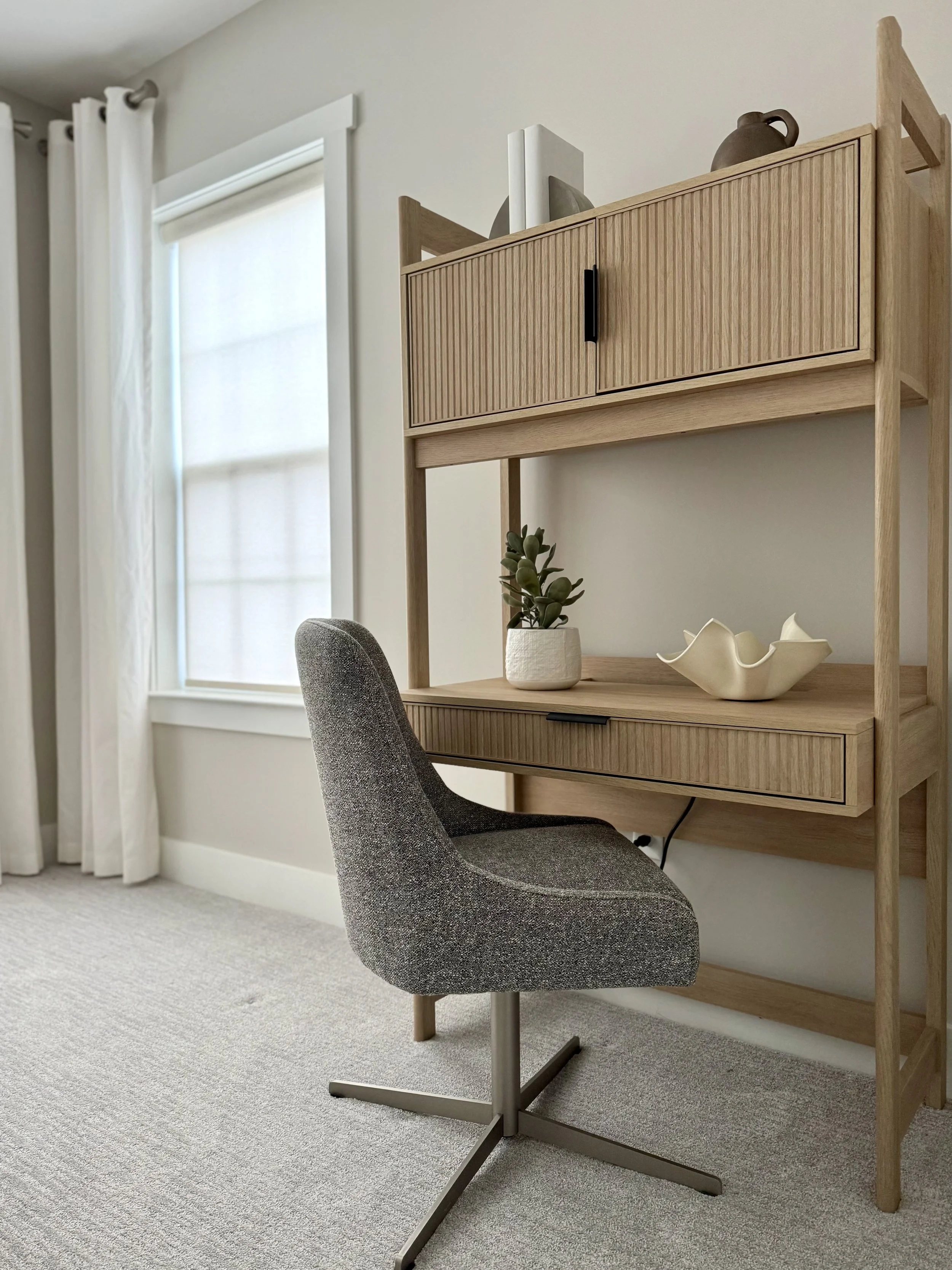 Modern minimalist home office with a gray swivel chair, wooden desk with built-in shelf, and decorative items like a vase and plant. Room features a large window with white curtains.