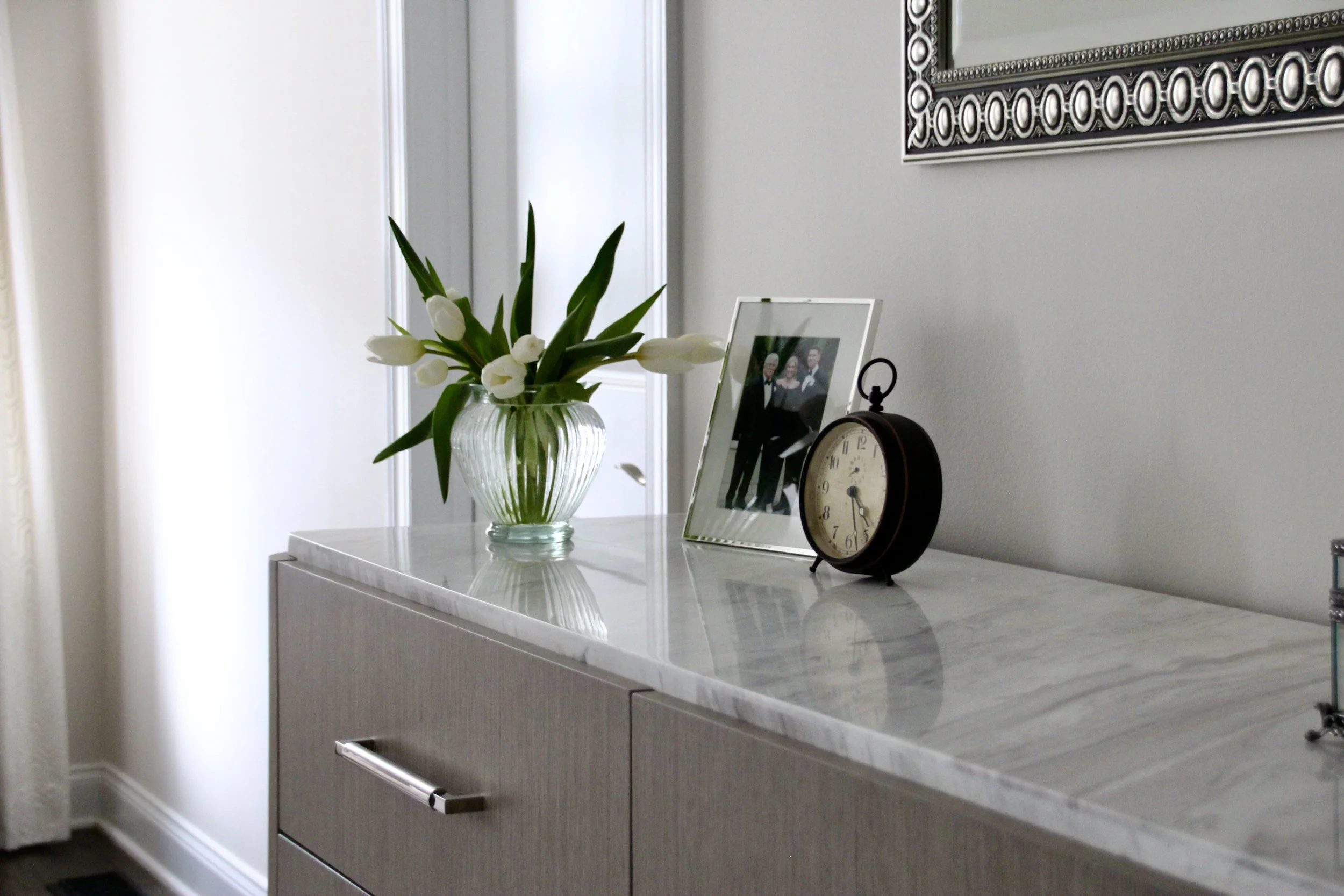 Vase with white tulips, framed photo, and vintage clock on marble-topped sideboard.