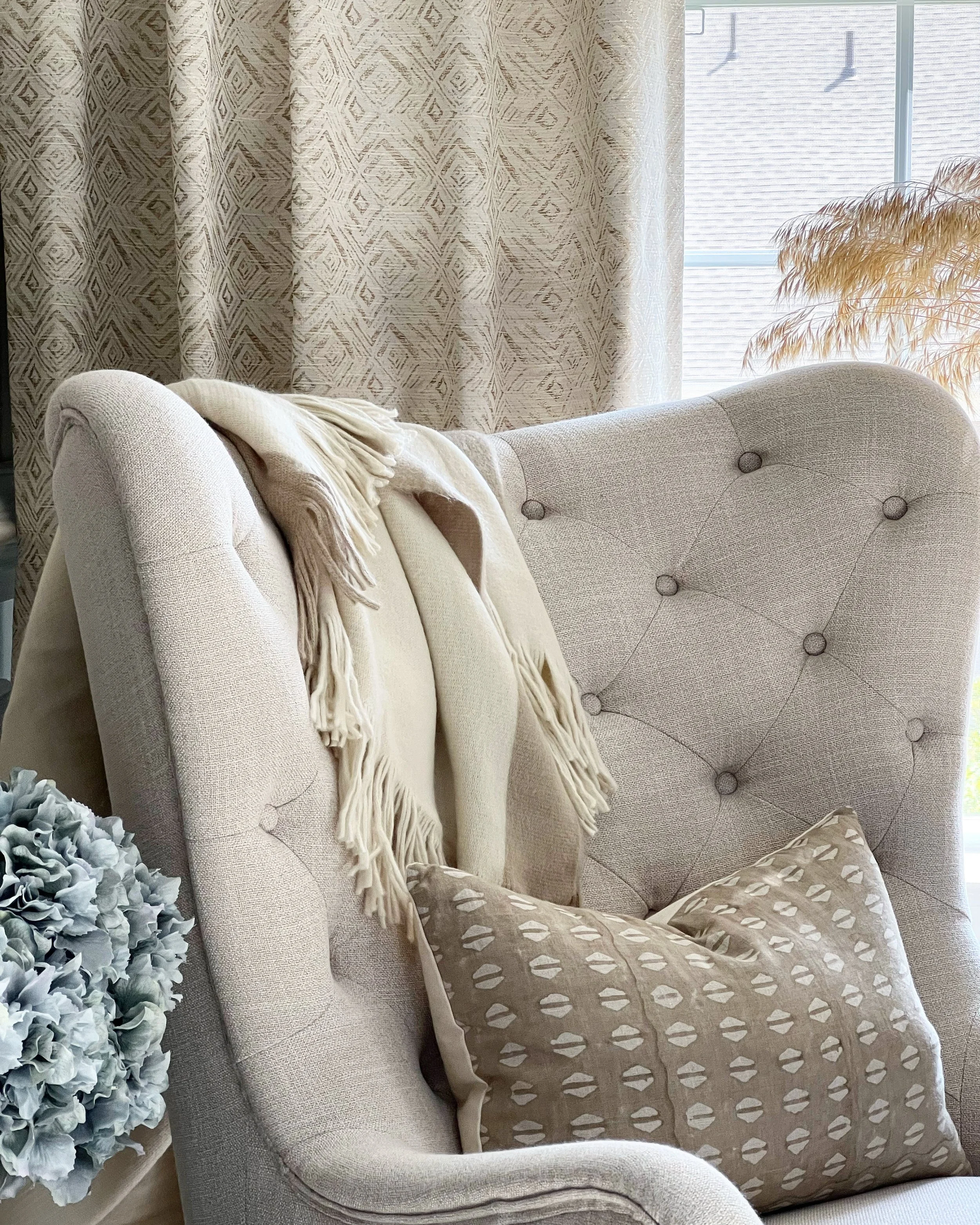 Beige tufted chair with a fringed throw blanket, patterned pillow, decorative plants, and patterned curtains.