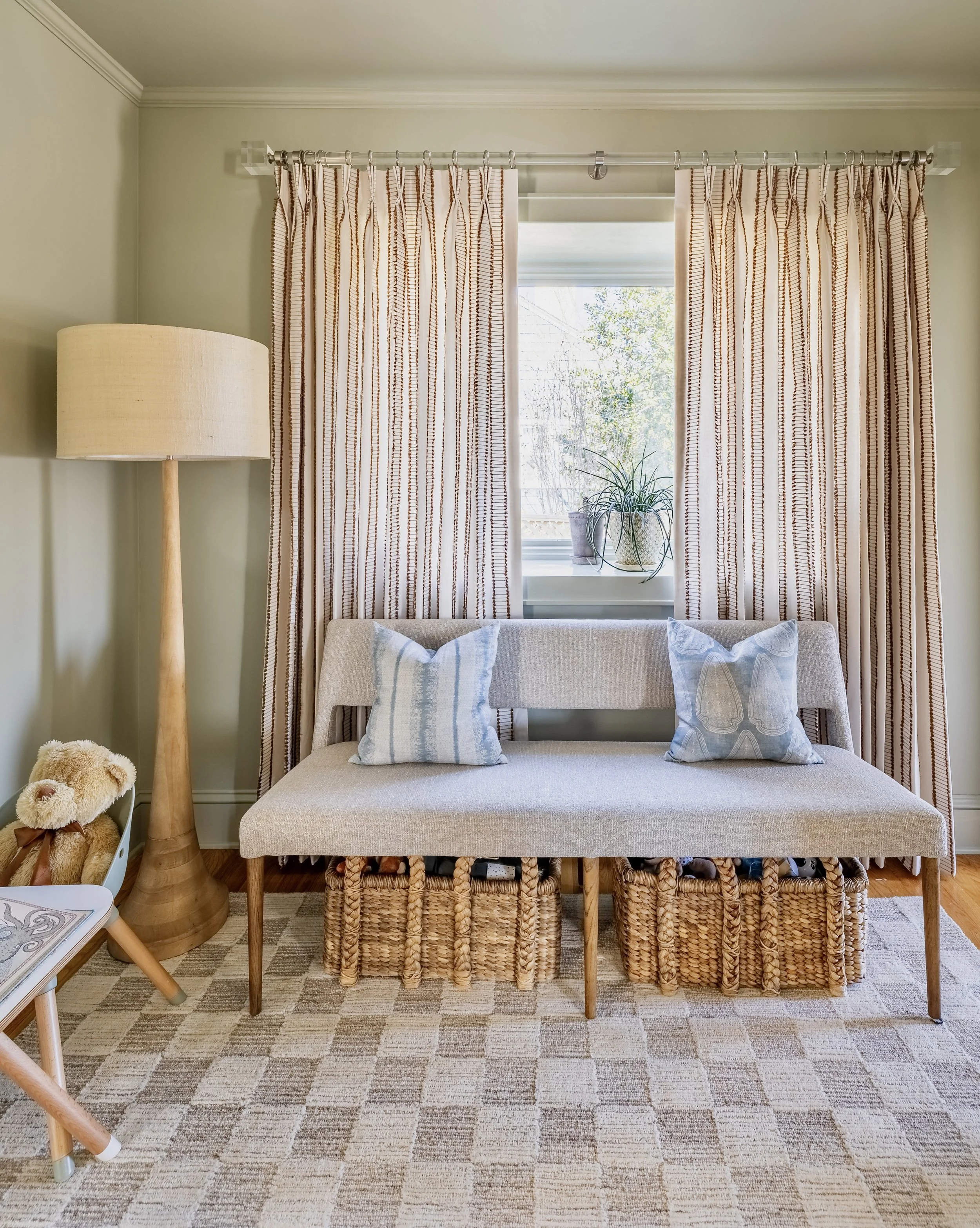 Living room with modern decor, featuring a light gray bench with blue pillows, wicker baskets underneath, a large floor lamp, patterned curtains, a window with potted plants, teddy bear on a side table, and a checkered rug.