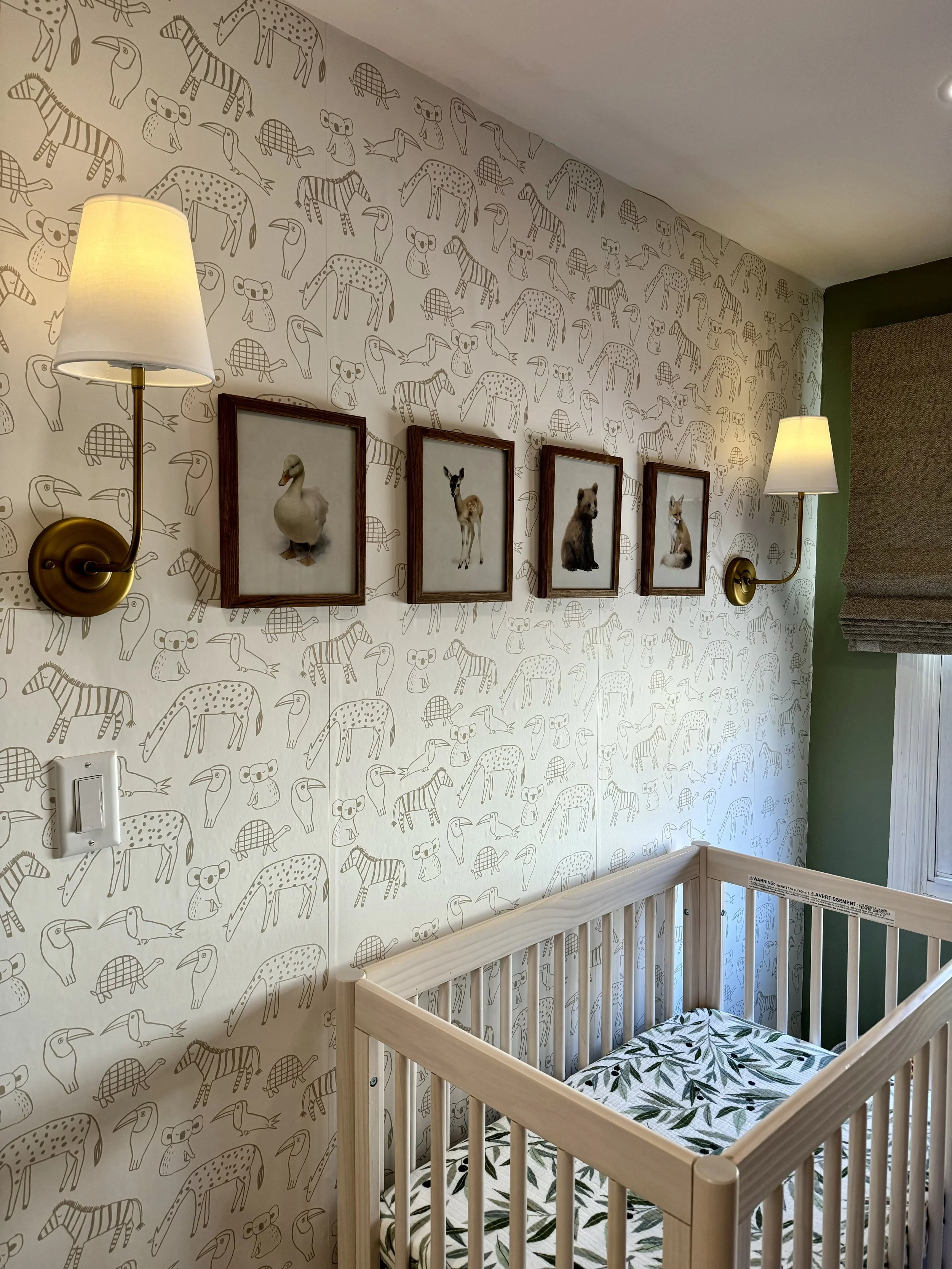A nursery room with animal-themed wallpaper, featuring stylized drawings of various animals. There are framed animal pictures on the wall above a wooden crib with a leaf-patterned mattress. Two wall-mounted lamps with white shades are on either side 