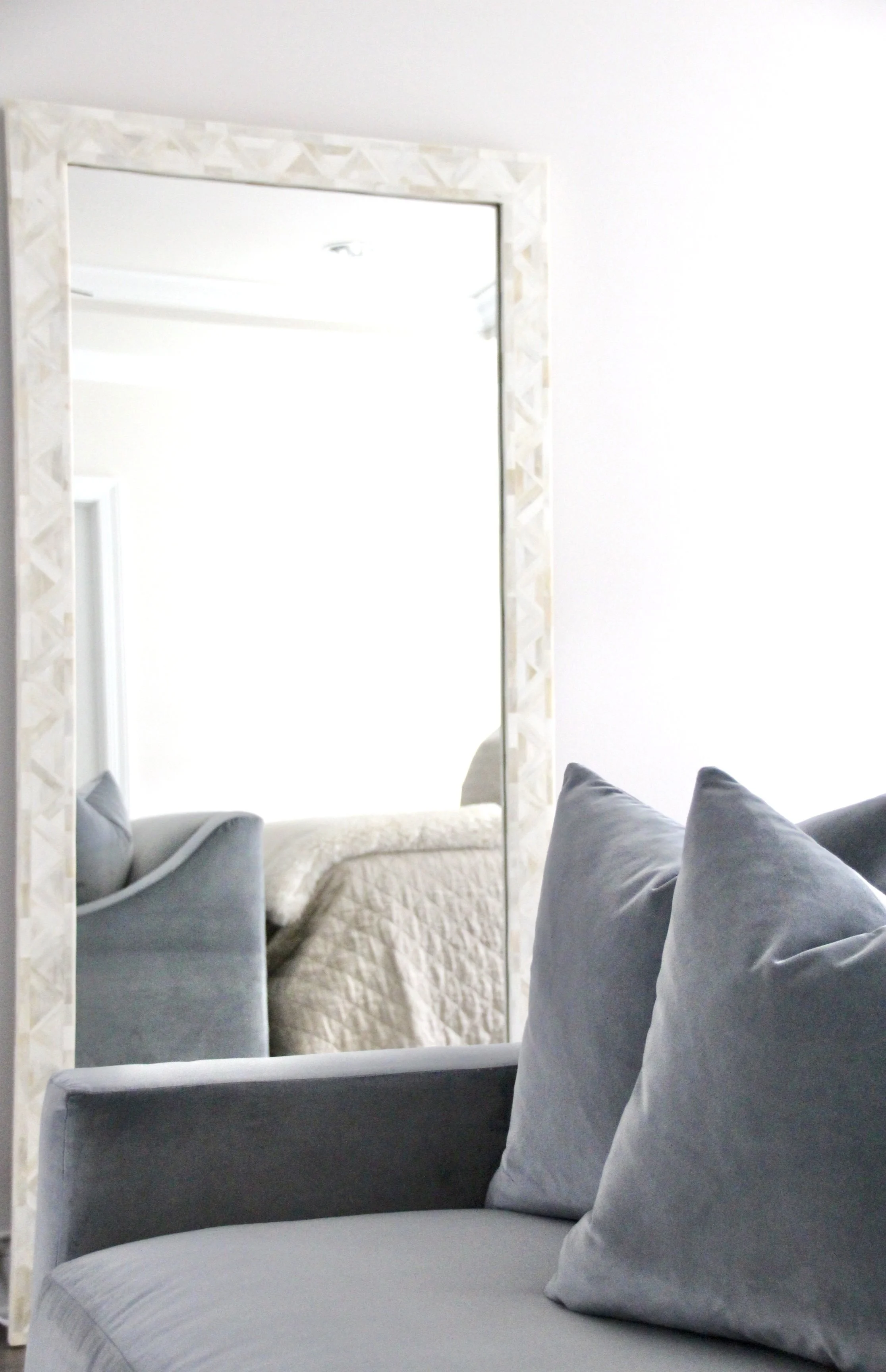 Mirror reflecting a gray sofa with pillows and a quilted bed in a bright room