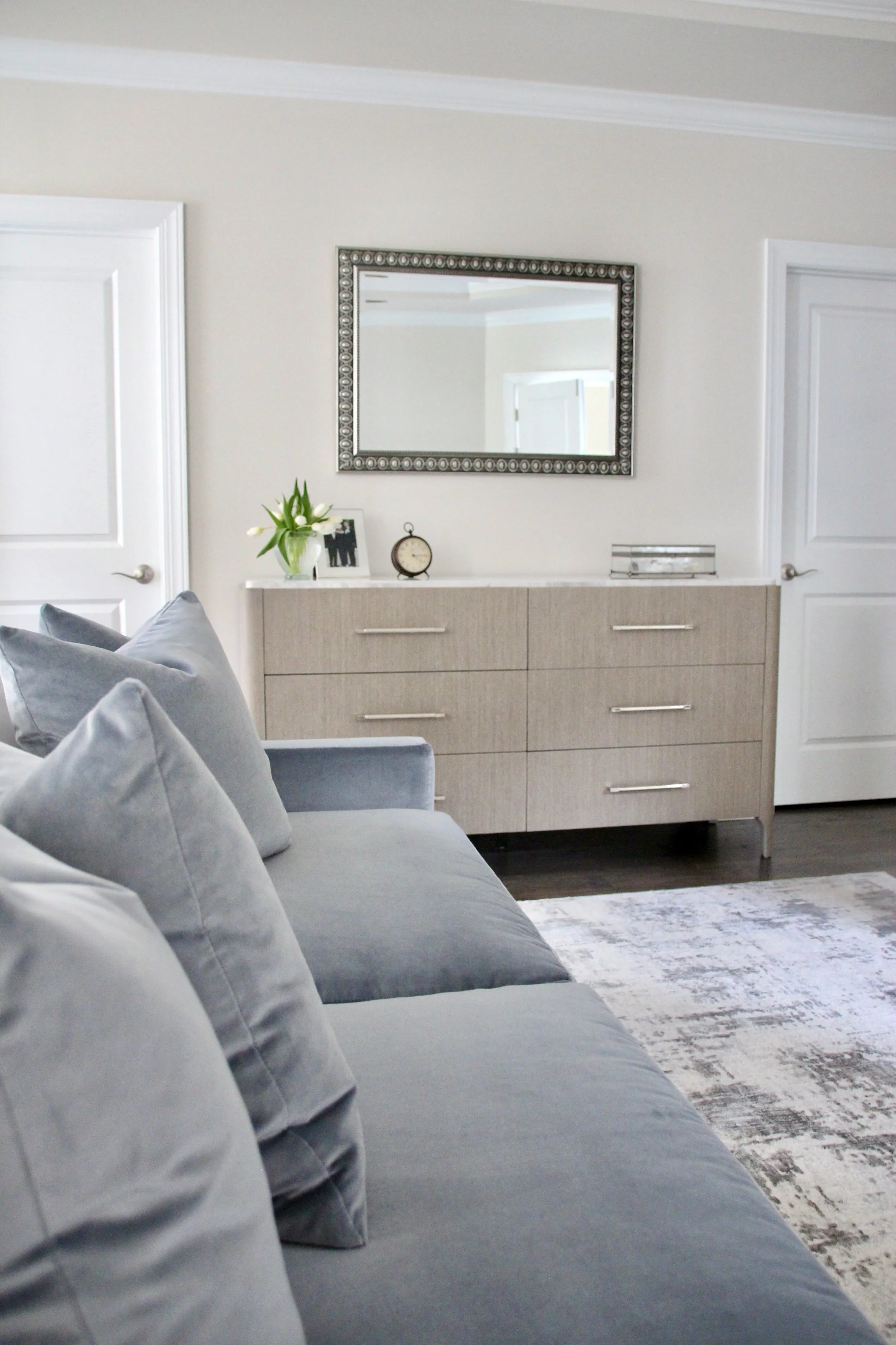 Stylish living room with a light gray sofa, decorative pillows, a wooden dresser with drawers, a wall mirror, and a vase with flowers.