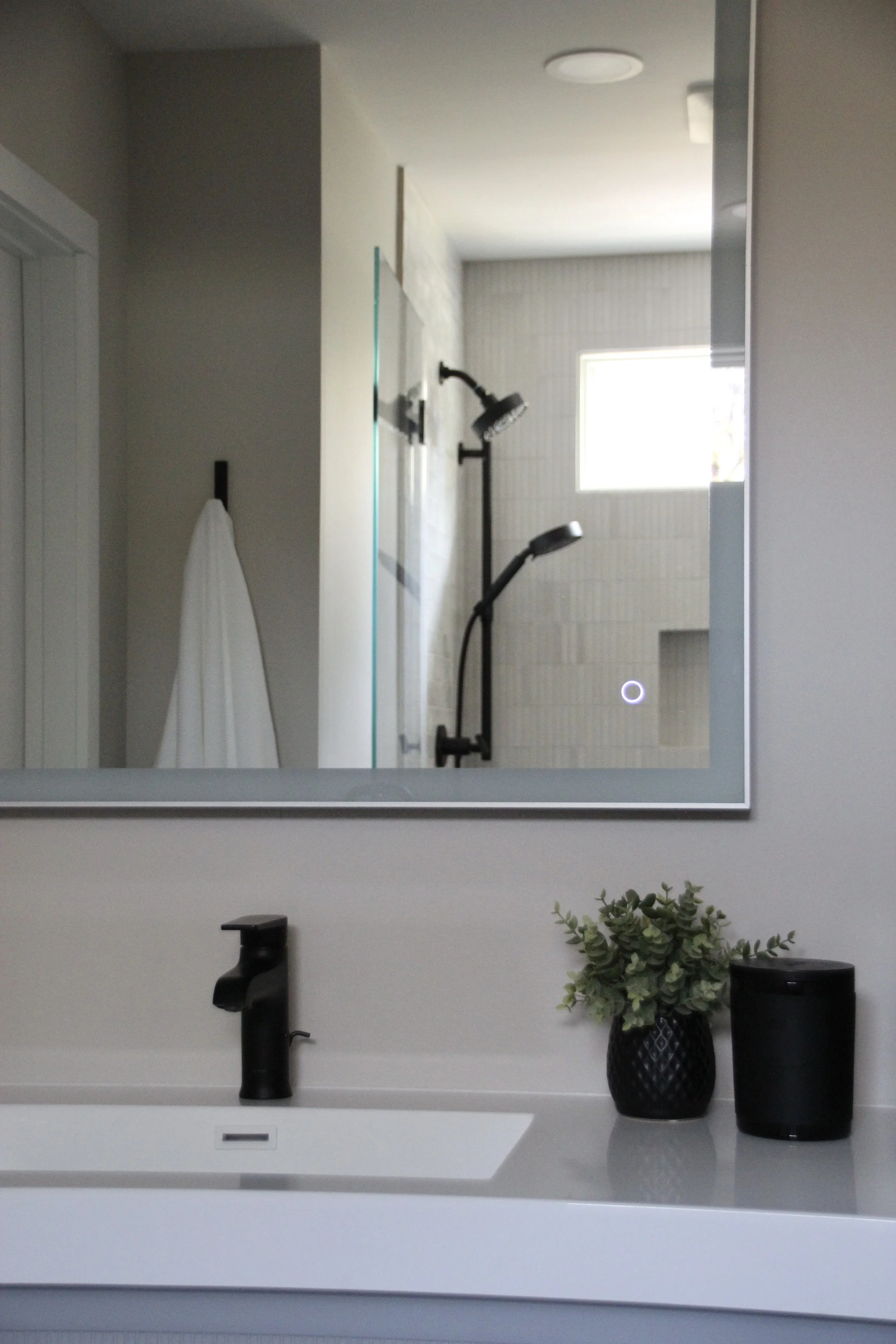 Modern bathroom with mirror reflection, black faucet, and decorative plants on countertop, showing shower area with black fixtures.