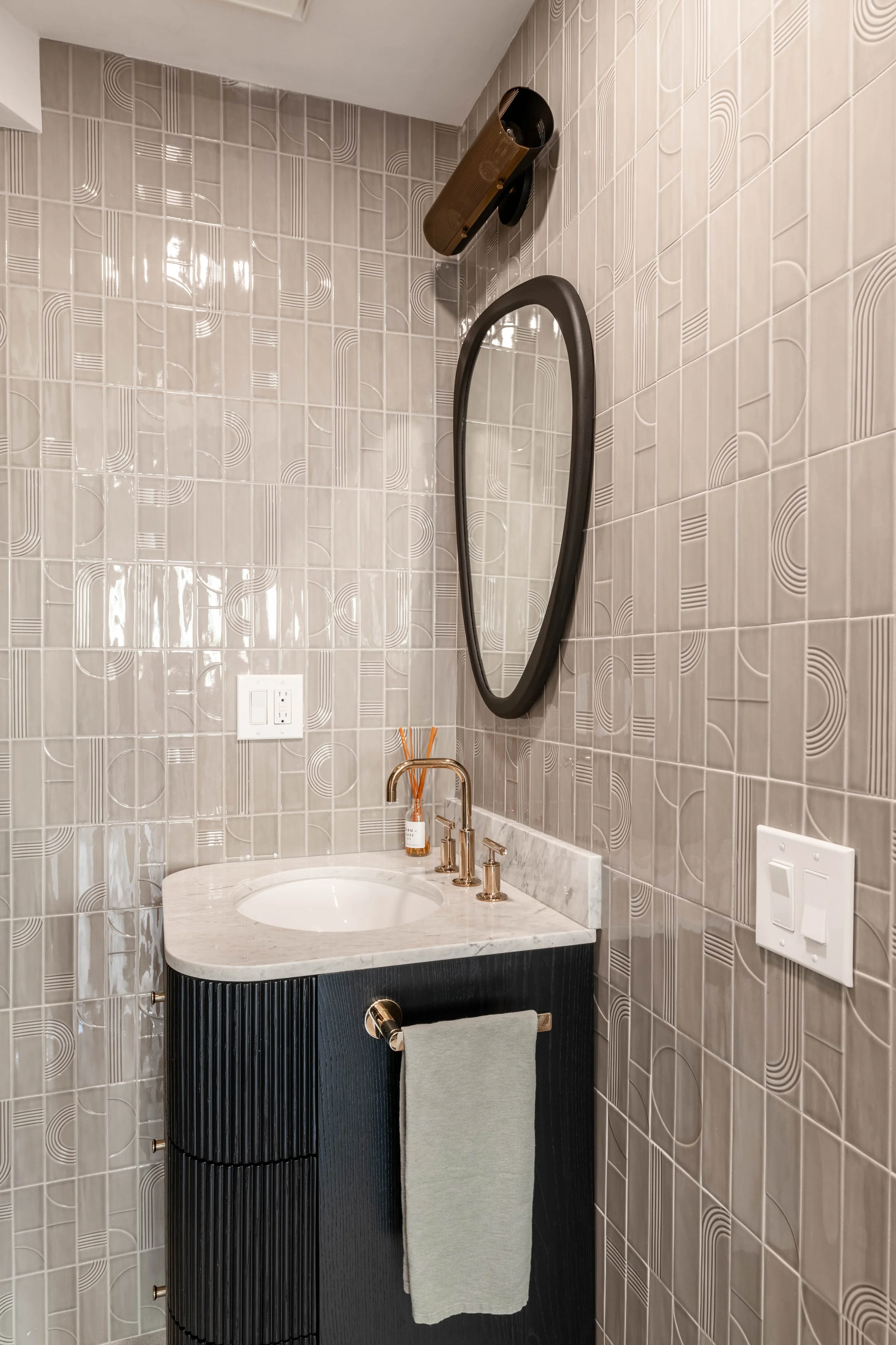Modern bathroom with geometric patterned tiles, oval mirror, marble countertop, gold faucet, towel rack with gray towel, and light switches.