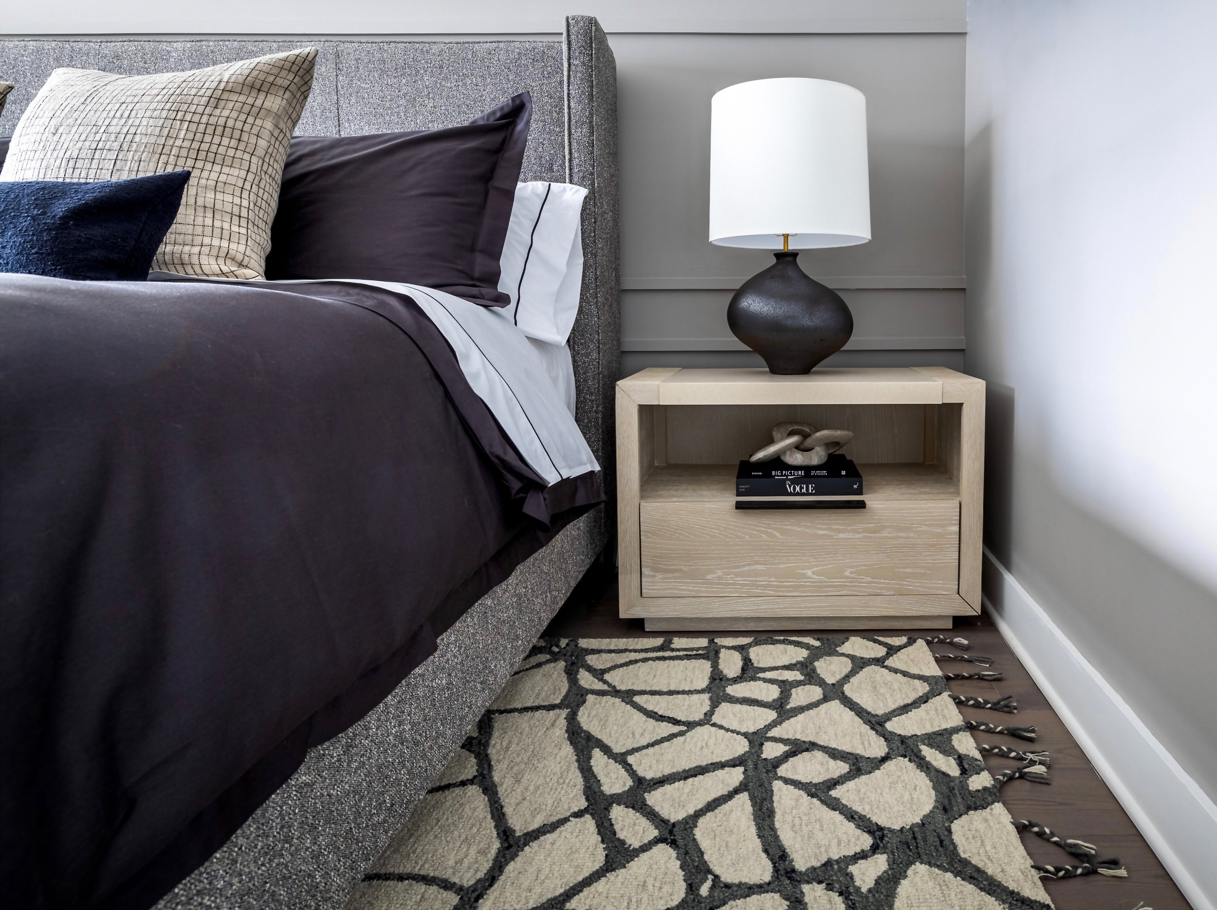Modern bedroom with grey upholstered bed, black and beige pillows, wooden nightstand, black lamp, and patterned rug.