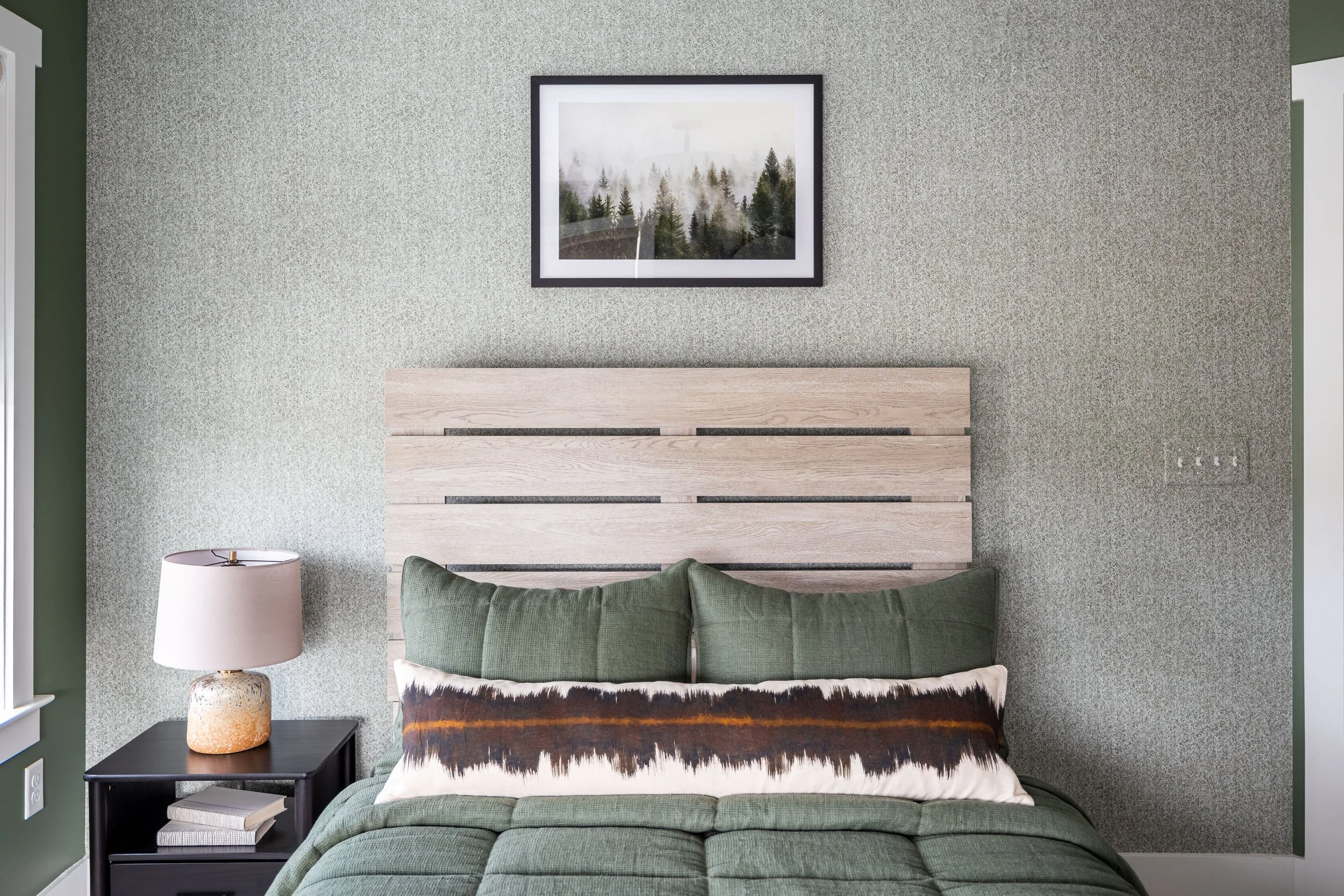 A modern bedroom with a green color scheme. The room features a wooden headboard, a green bedspread, and a decorative pillow with a nature-inspired design. A bedside table holds a lamp and books. On the wall above the bed, there's a framed picture of