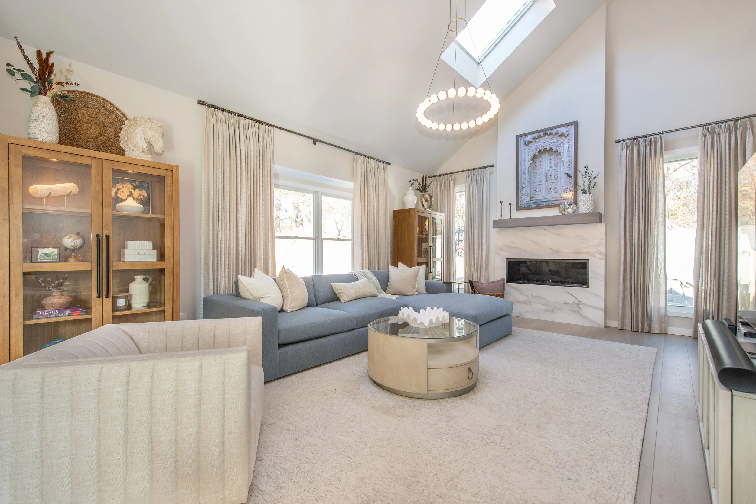 Modern living room with a blue sectional sofa, beige armchair, glass coffee table, and light curtains. Features a fireplace with marble surround, skylight, chandelier, and decorative shelves.