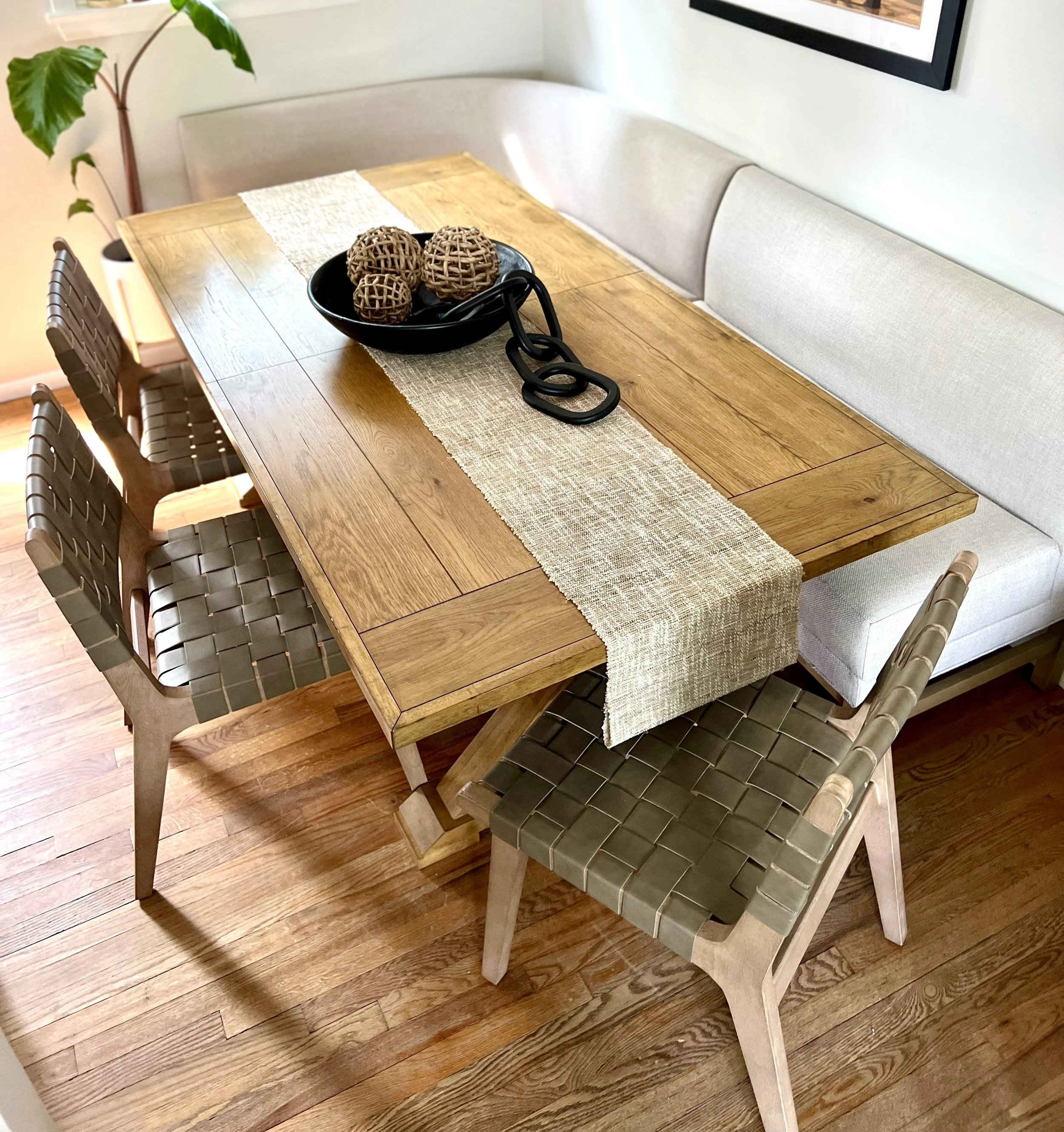 A wooden dining table with an off-white runner and decorative items in a black bowl. The table is surrounded by woven chairs and a light-colored bench. A houseplant is visible in the corner of the room.