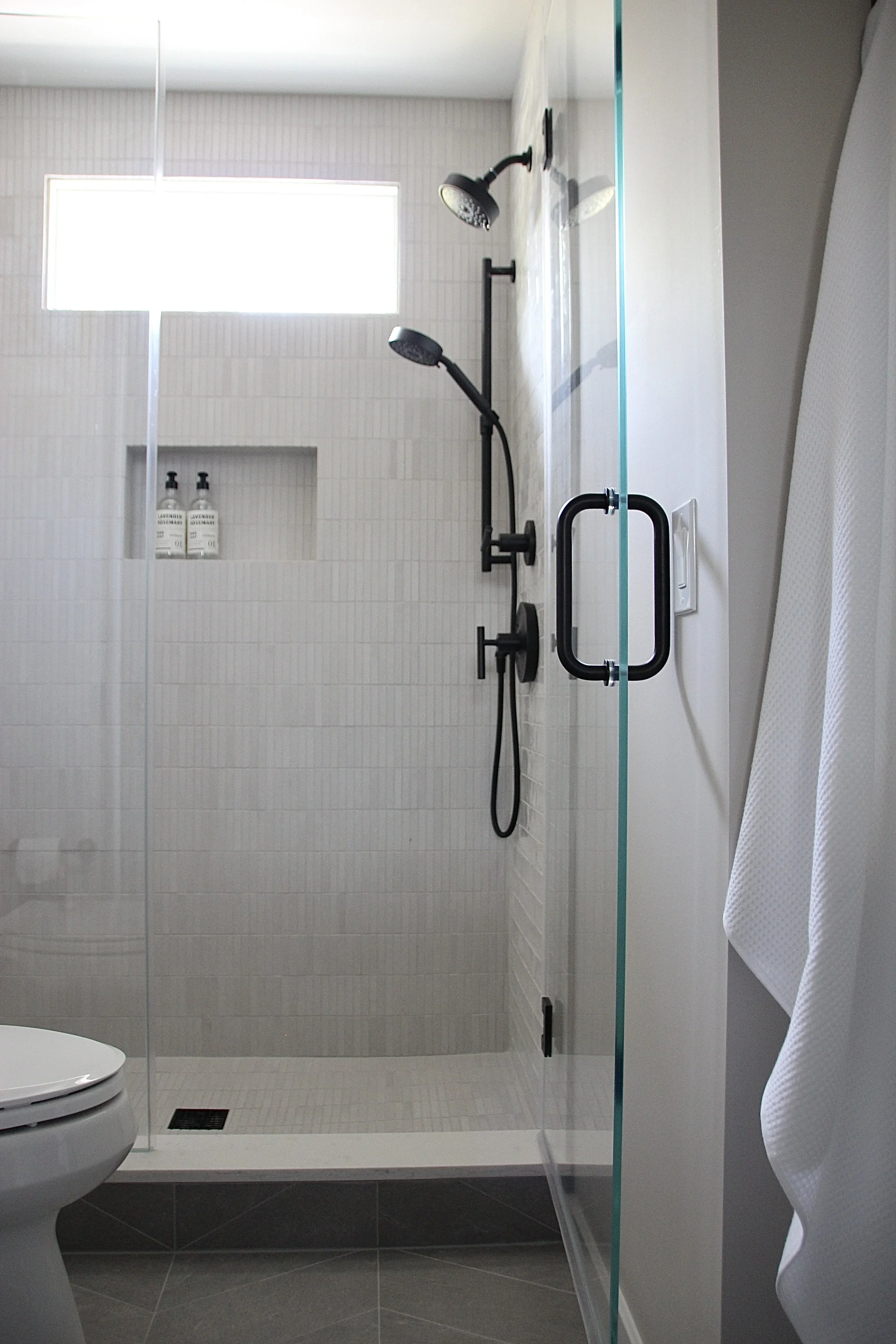 Modern bathroom with glass shower enclosure, black fixtures, and white tiled walls. A towel hangs on a hook outside the shower.