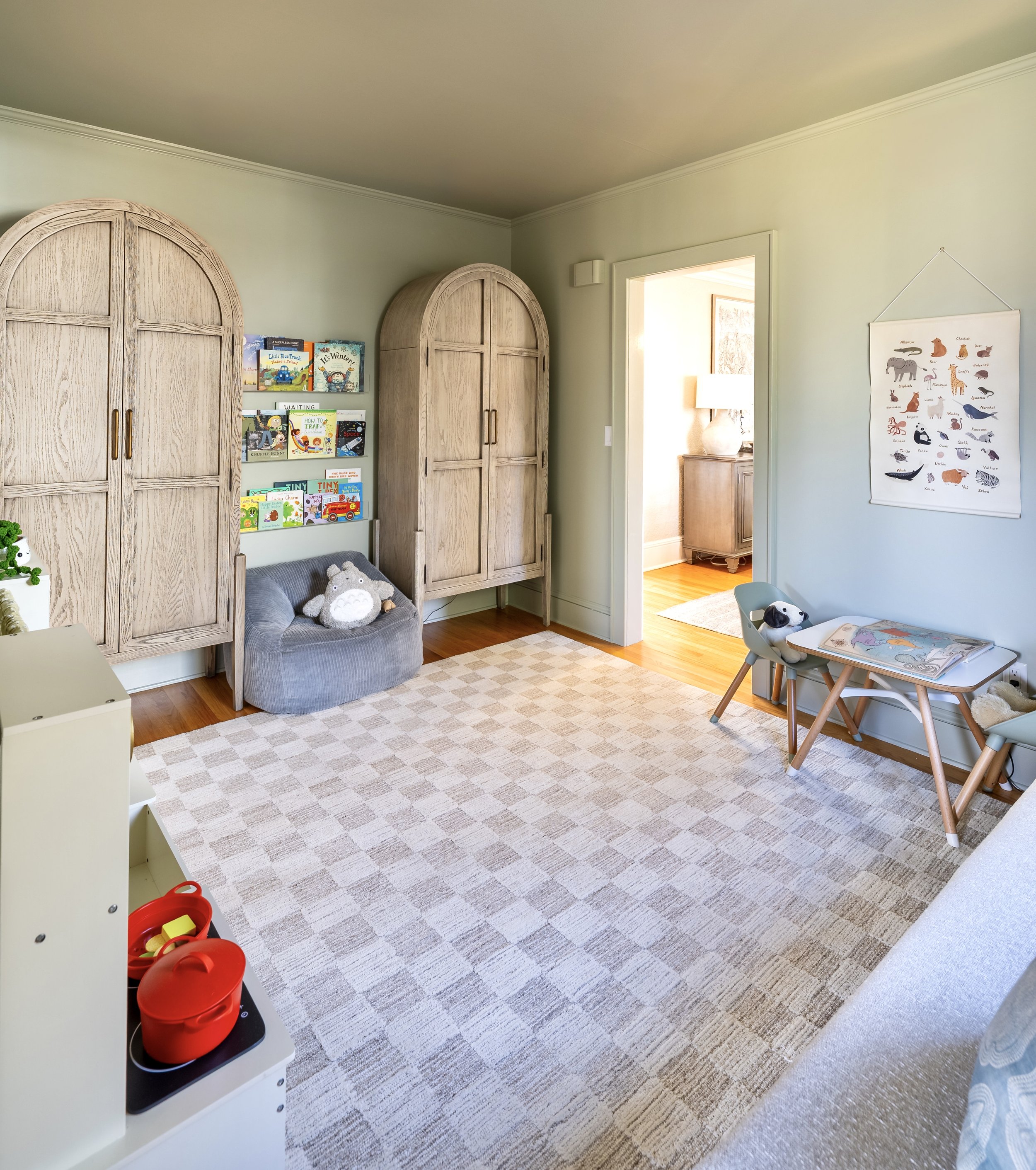 A child's playroom featuring a checkered rug, two arched wooden cabinets, and a small reading area with books displayed on shelves. A cozy chair with stuffed toys is in the corner. A small table and chair set is against the wall, with a poster displa