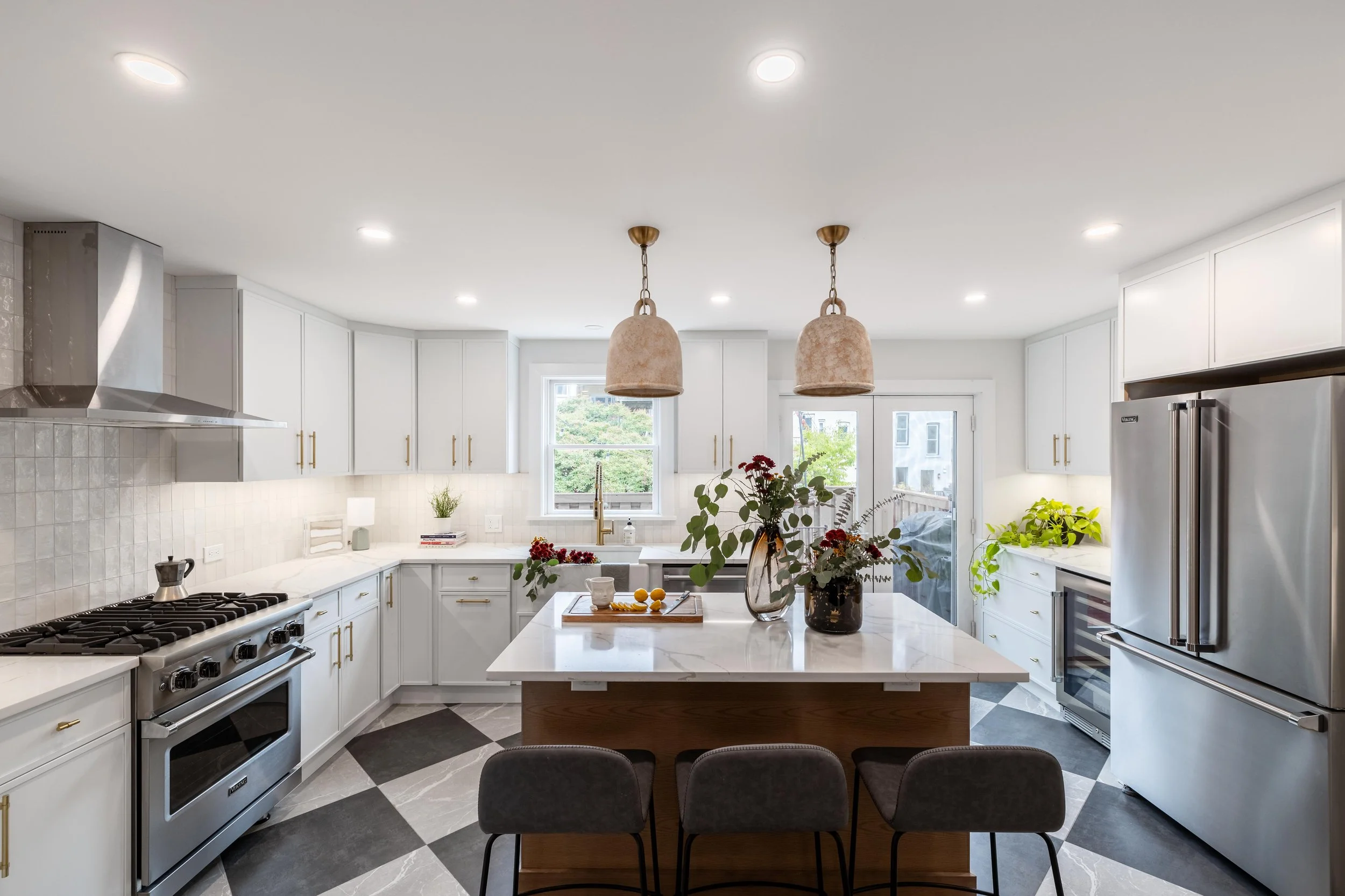 Modern kitchen with stainless steel appliances, white cabinetry, marble countertops, island with barstools, pendant lighting, and indoor plants.