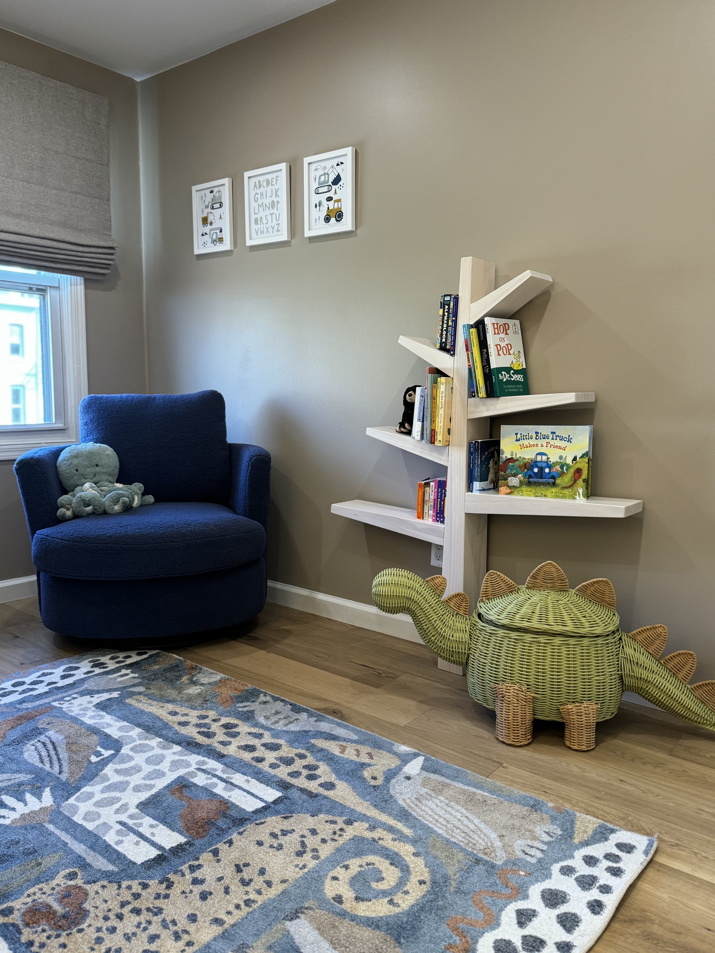 Cozy children's room with a blue chair, stuffed animal, wooden bookshelf shaped like a tree holding books, dinosaur-themed wicker stool, and a decorative animal print rug.