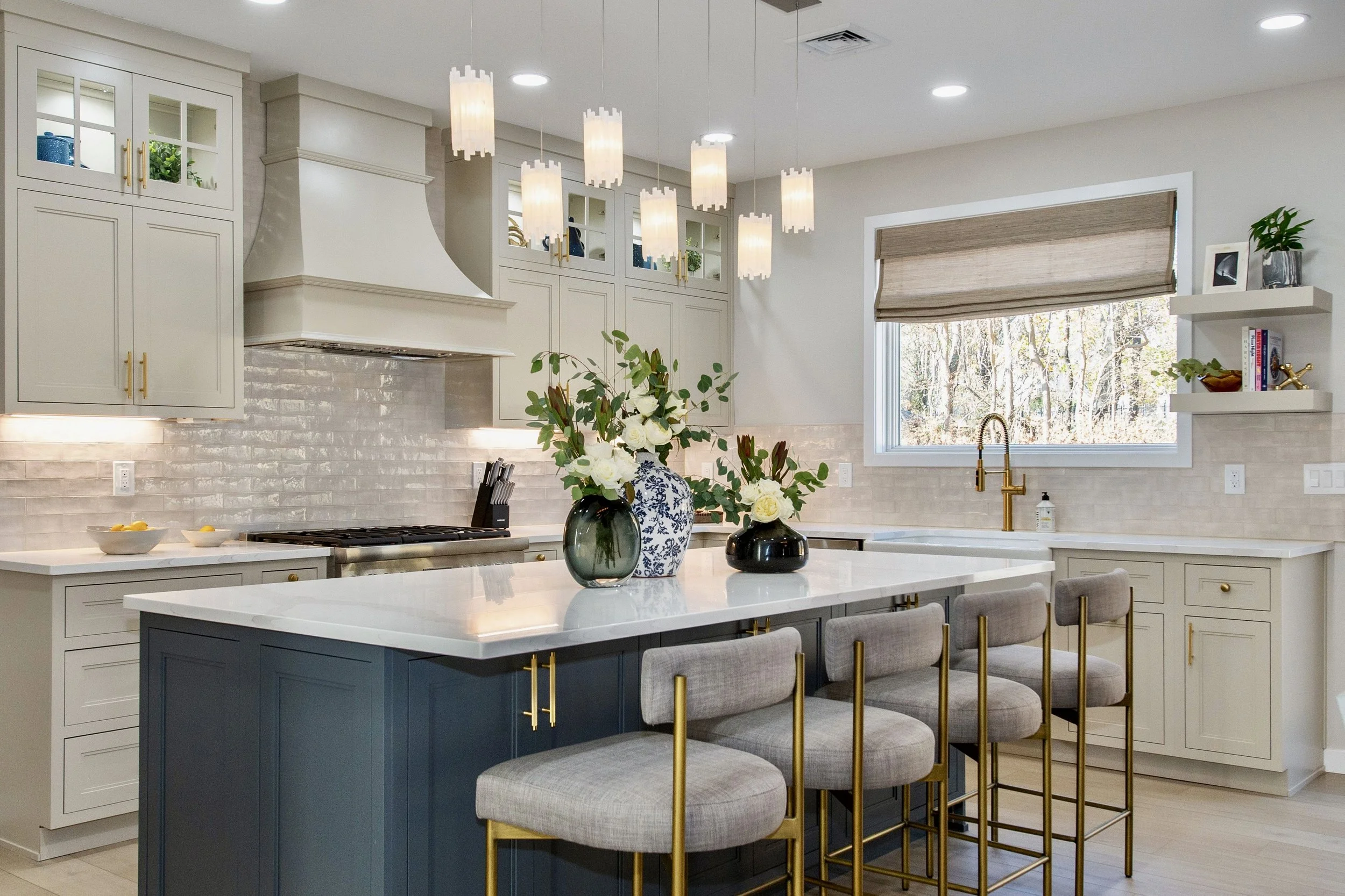 Modern kitchen with a center island, bar stools, pendant lighting, light cabinets, and a window. Decor includes vases with flowers on the island and wall shelves with books and decor items.