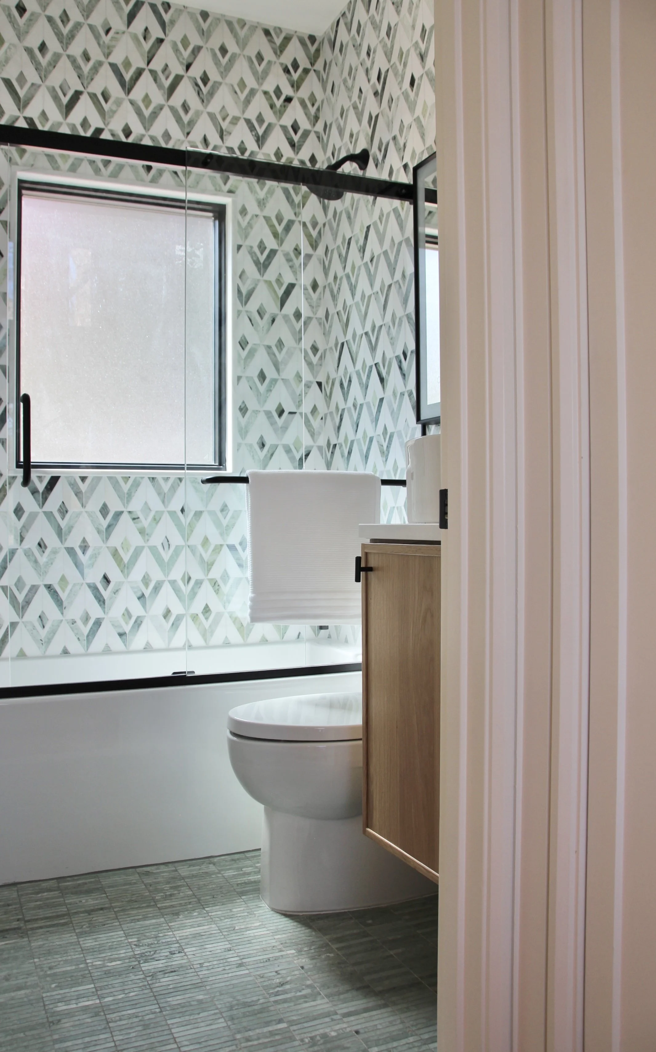 Modern bathroom with patterned tile wall, bathtub, toilet, and wooden vanity.