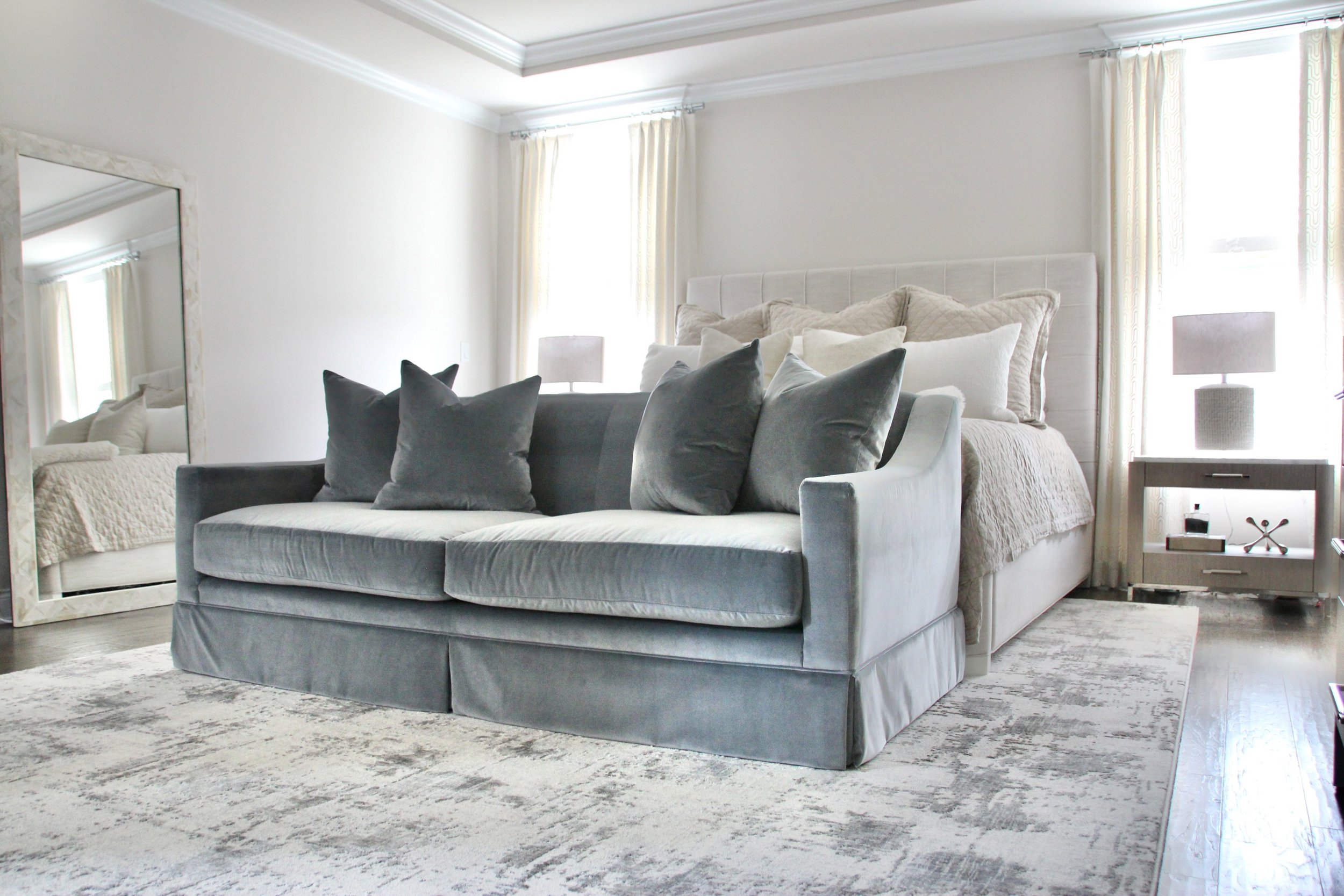Modern bedroom with a gray velvet couch at the foot of a bed, large mirror, bedside lamps, and light curtains.