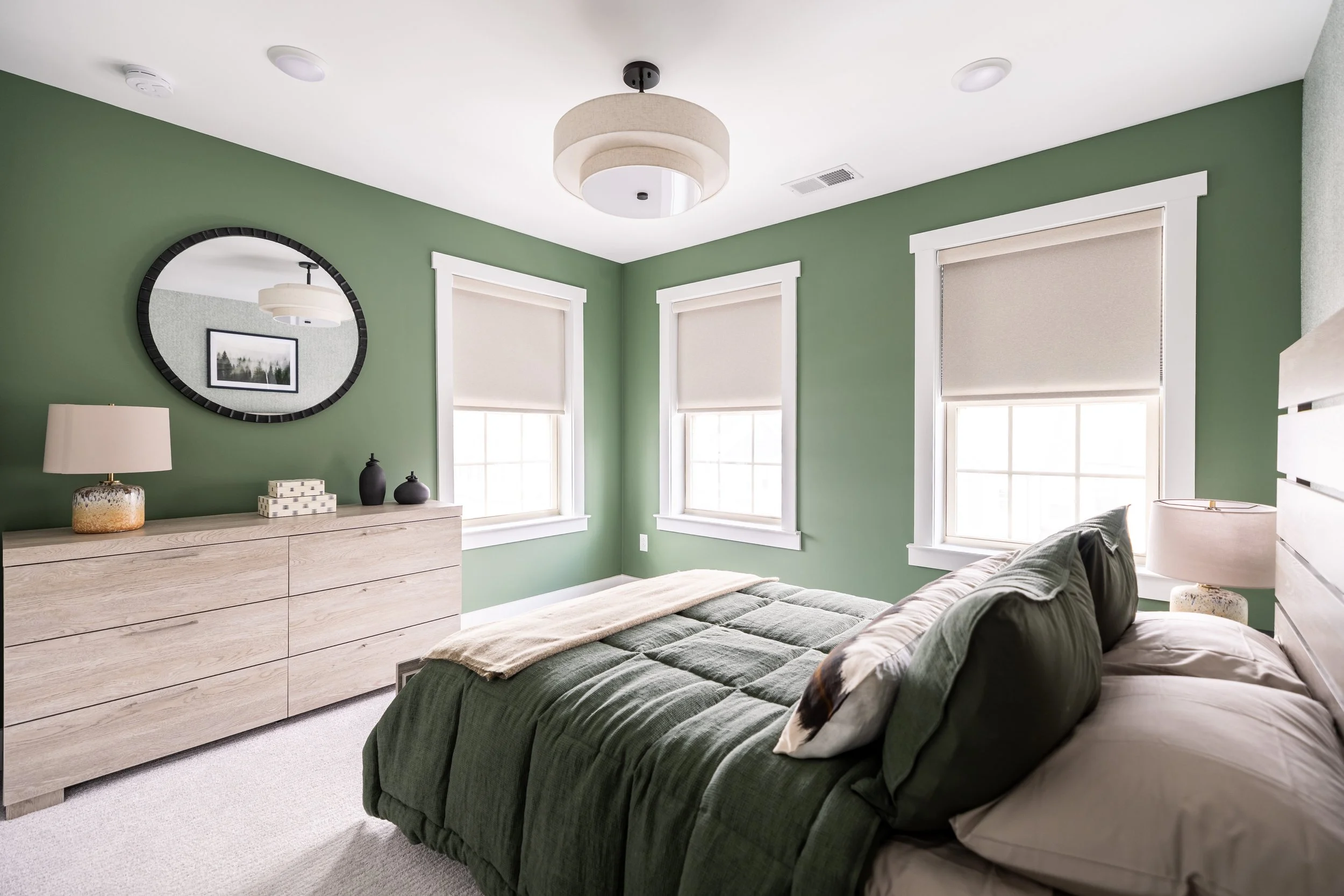 Modern bedroom with green walls, beige carpeting, light wood dresser, round mirror, ceiling light, green bedding, and beige window shades.