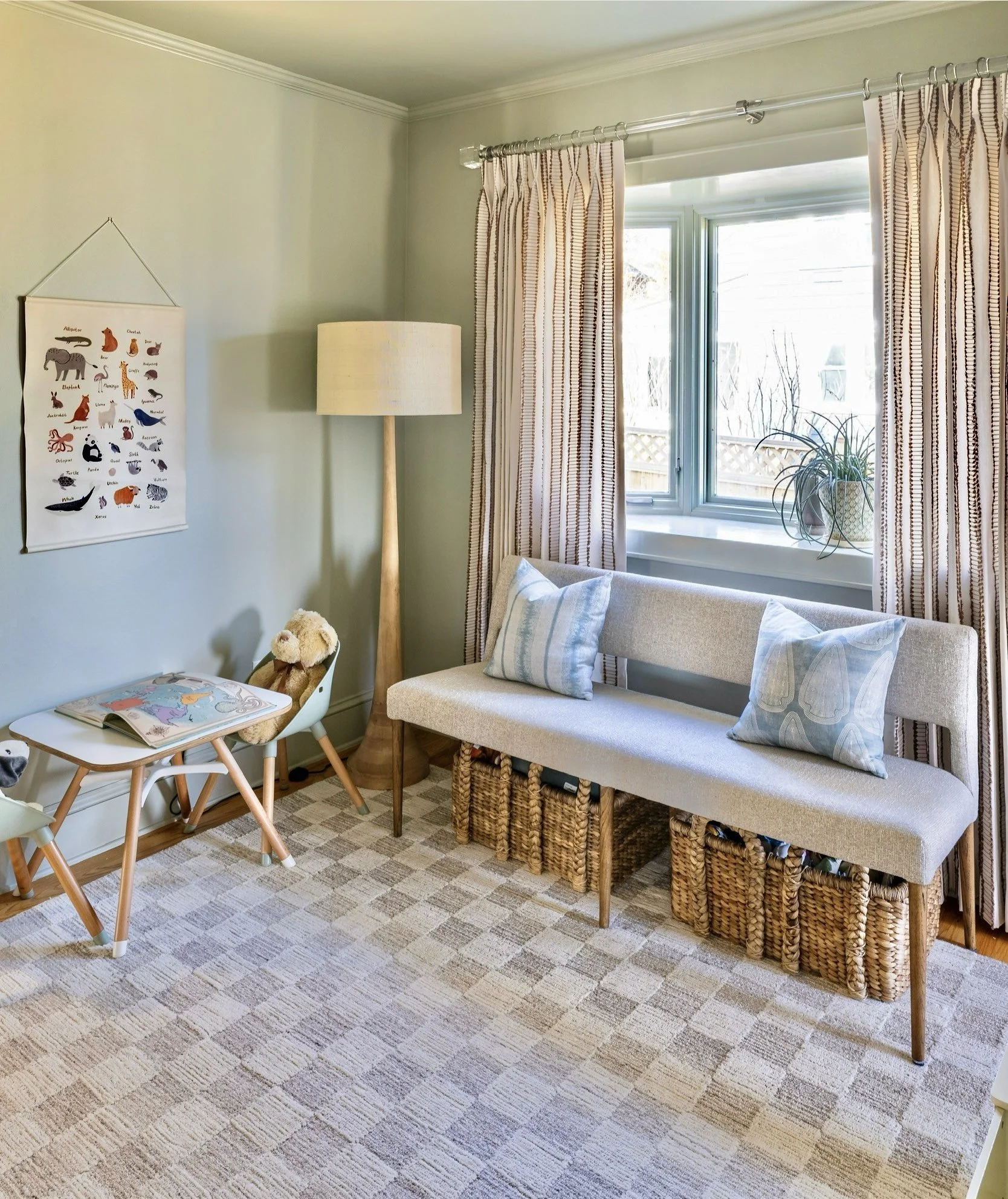 Cozy living room corner with a gray bench, blue plaid pillows, and wicker baskets underneath. A light-colored rug covers the floor, and a round floor lamp stands next to a window with striped curtains. A white table holds a children's book beside a c