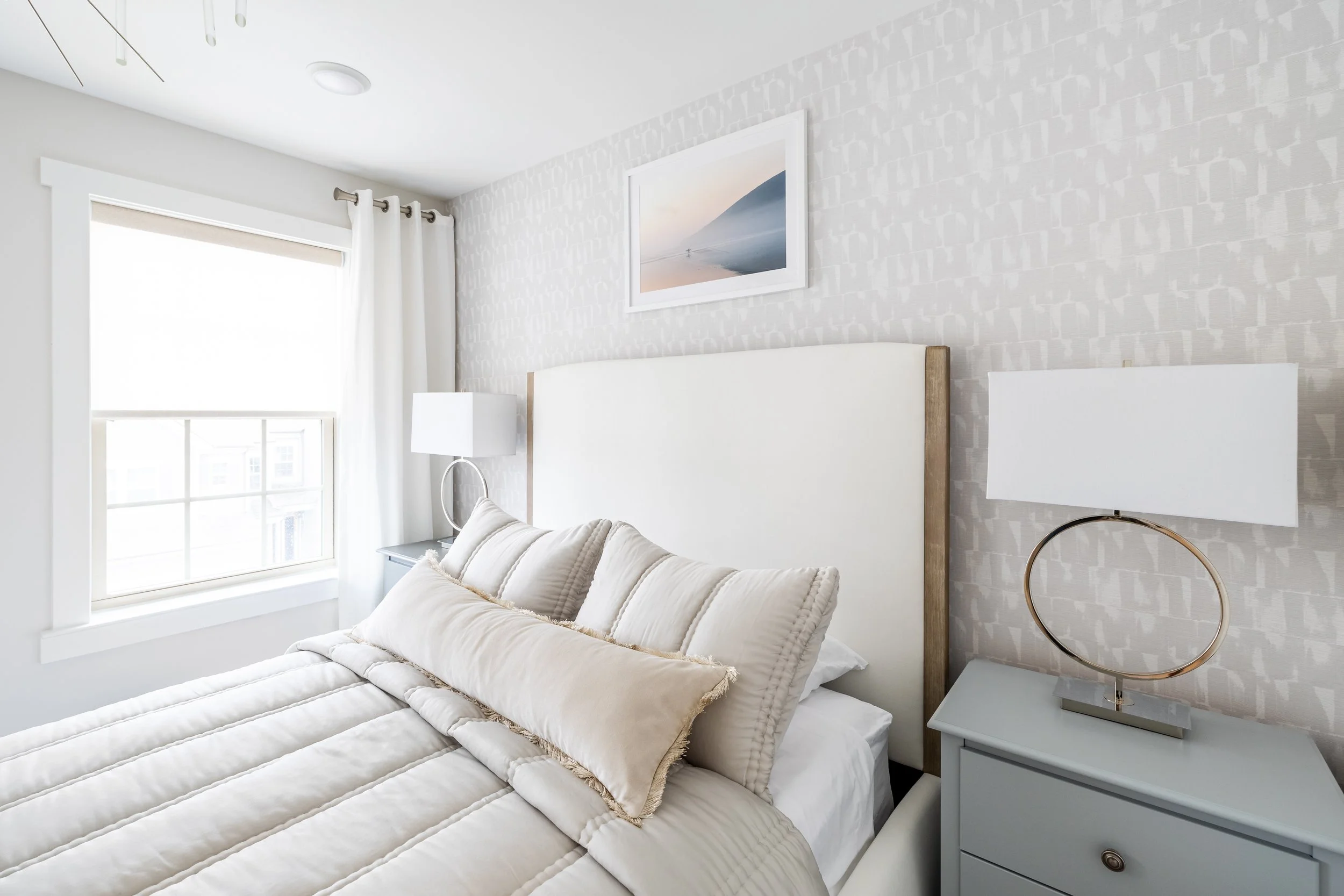 Modern bedroom with neutral decor, featuring a white upholstered bed, beige bedding, two bedside tables with lamps, and a framed landscape photograph.
