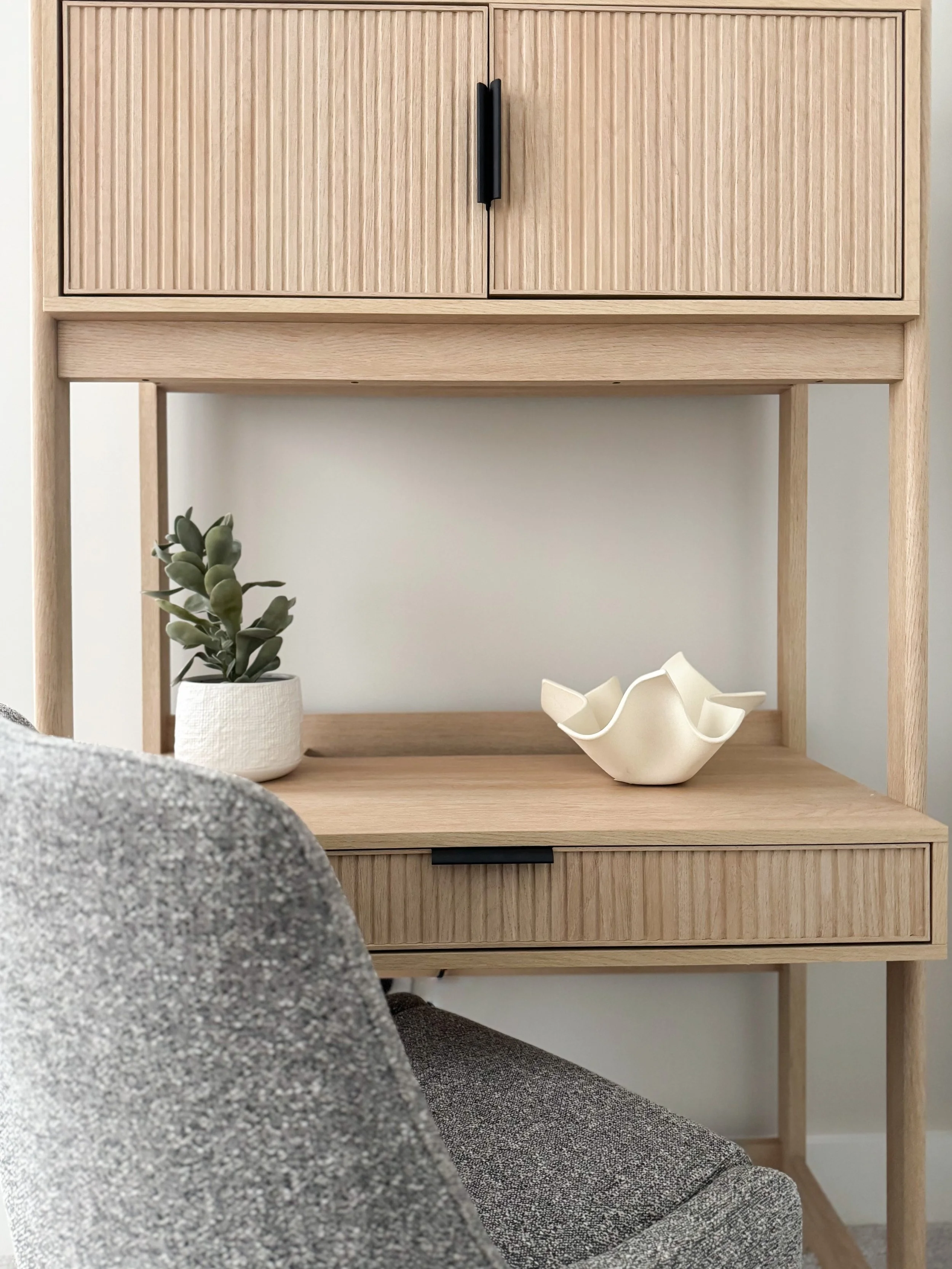 Minimalist desk with a decorative succulent plant in a white pot and a sculptural bowl on a wooden surface, accompanied by a grey upholstered chair.